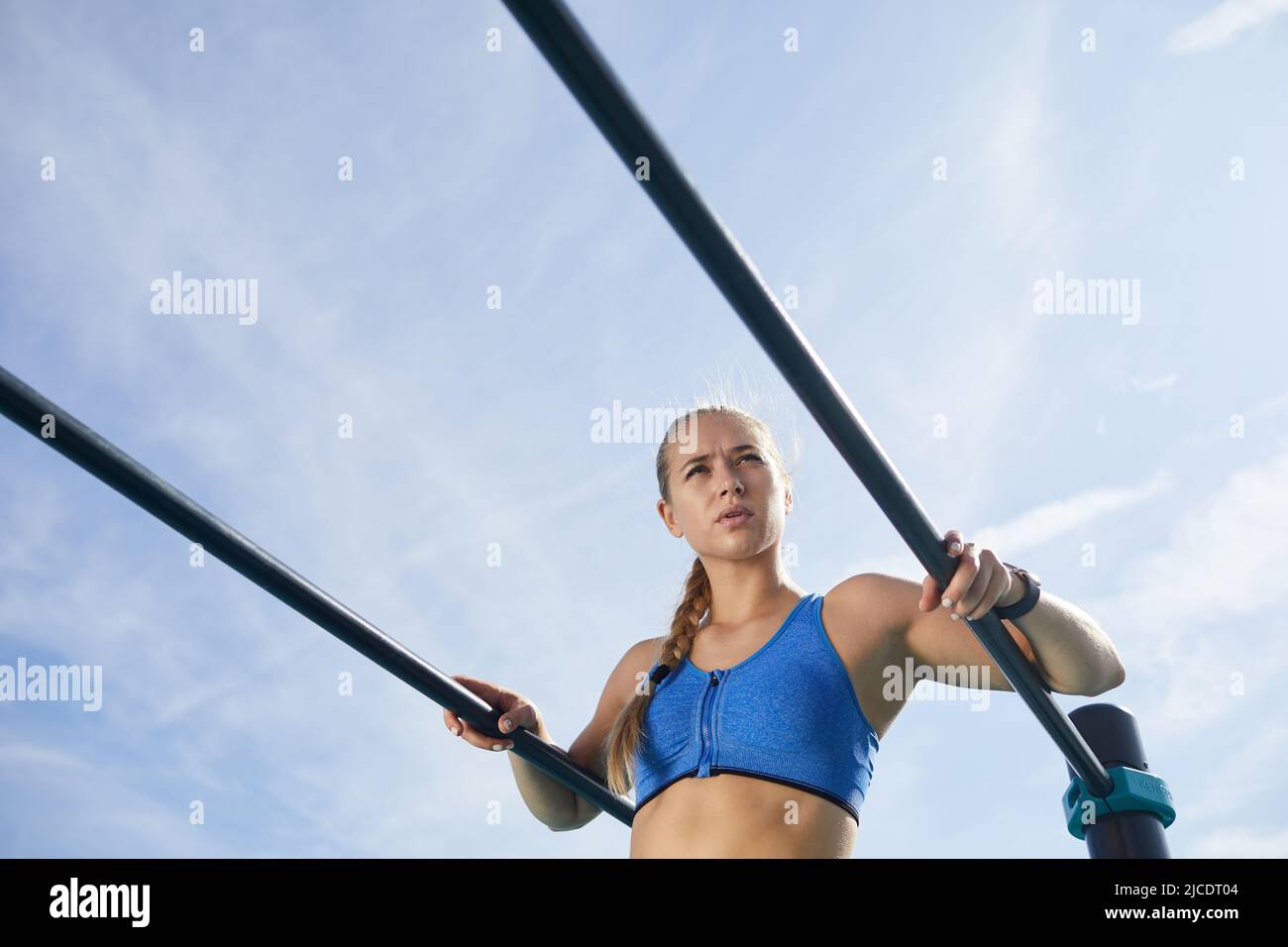 Giovane donna attraente e intenzionale con treccia di formazione su barre parallele all'aperto: Contempla intorno mentre prende la pausa tra le esercitazioni Foto Stock