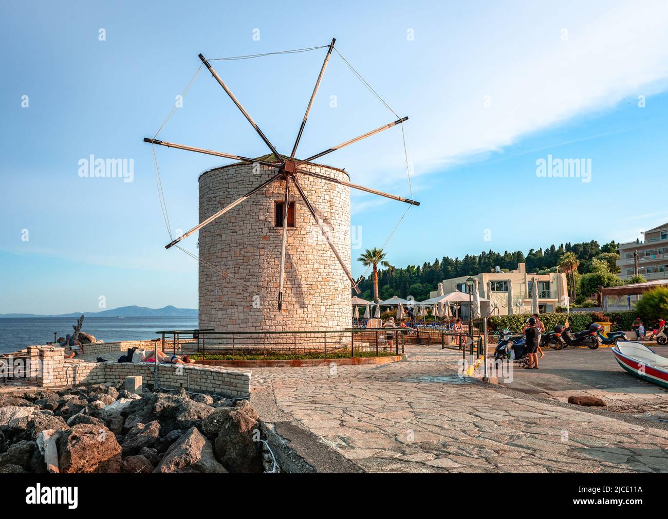 Corfù, Grecia - Giugno 3 2022: Il mare ricostruì vecchio mulino a vento tradizionale e la piccola passeggiata con ciottoli di pietra nel pomeriggio. Foto Stock
