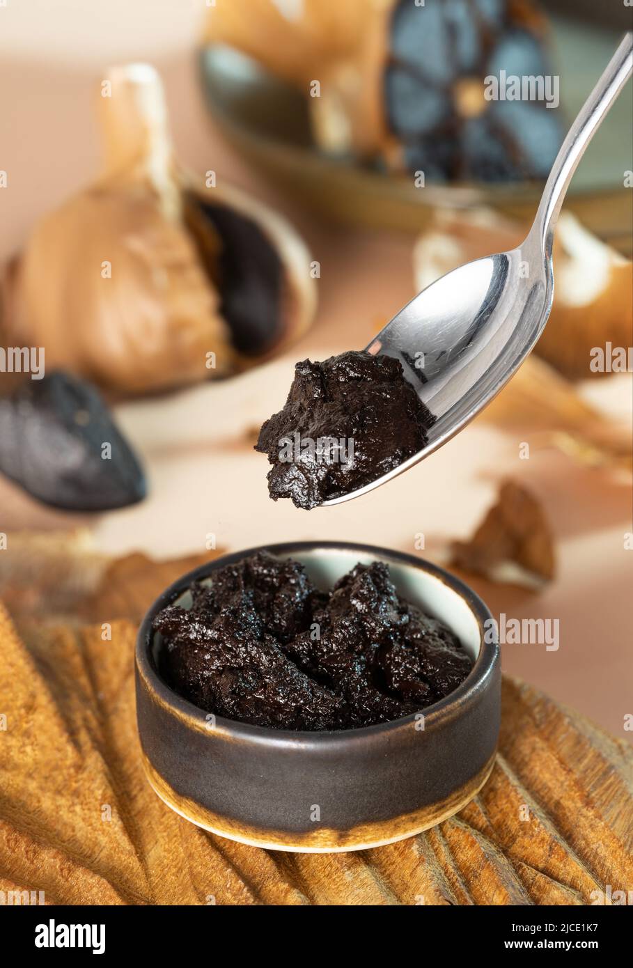 Cucchiaio con pasta d'aglio nera. Una piccola ciotola di argilla con aioli neri su un piatto di legno fatto a mano. Bulbo fermentato all'aglio sullo sfondo. Sano Foto Stock