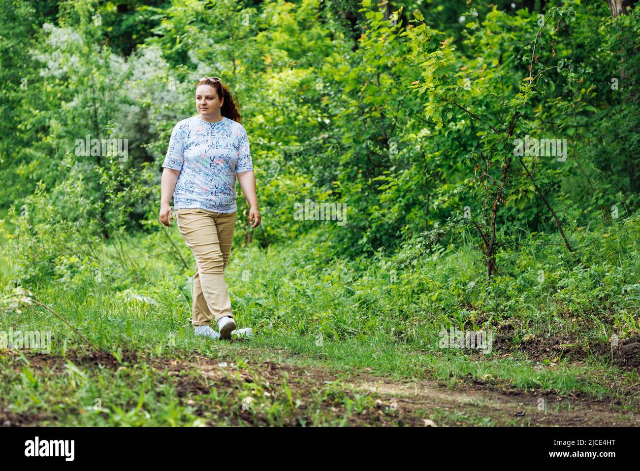 Ritratto di giovane donna grassa con lunghi capelli rossi ricci che camminano sul sentiero nella foresta del parco tra alberi verdi. Estate, fine settimana. Foto Stock