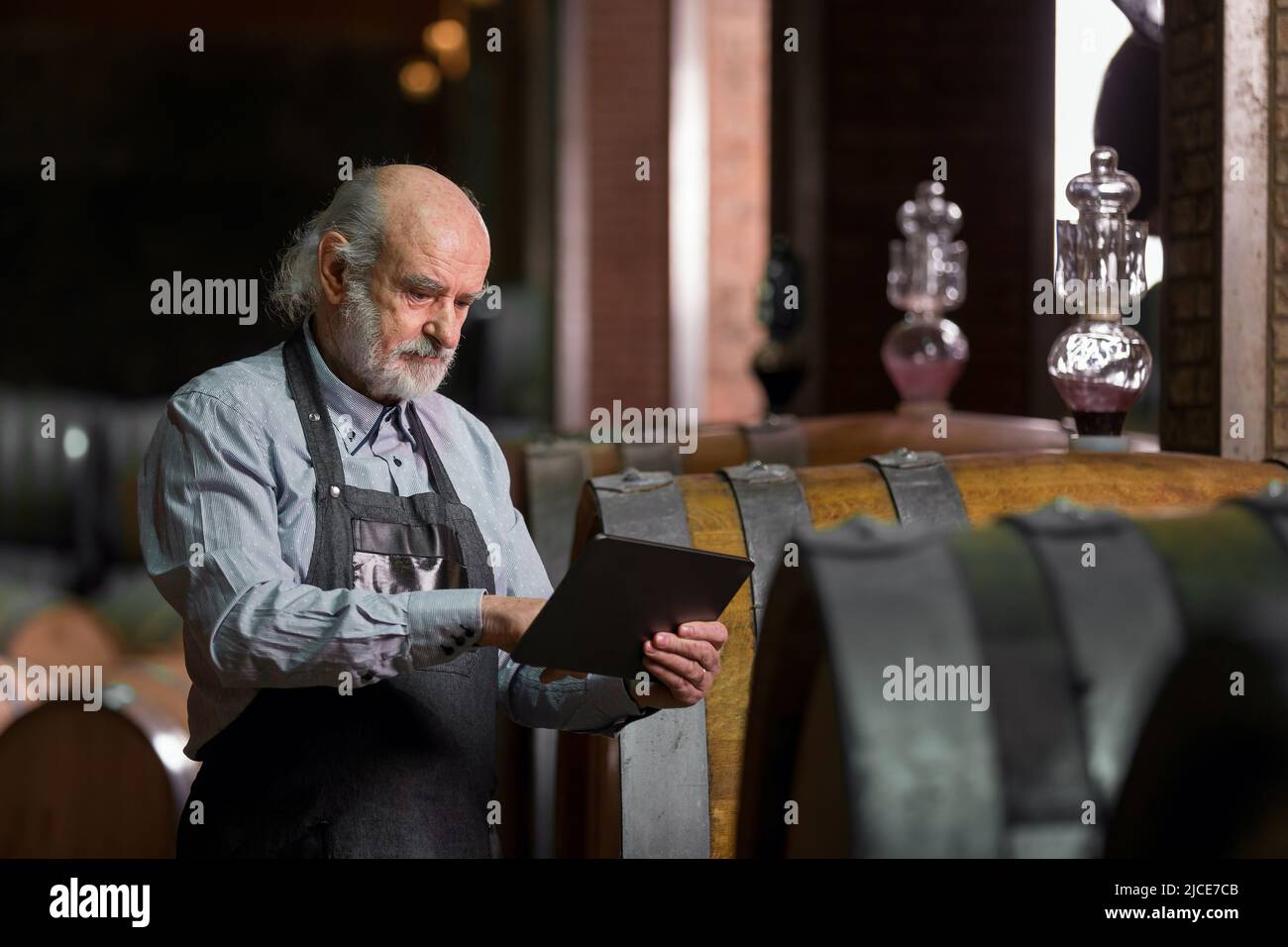 Uomo anziano caucasico, un enologo di greybeard con grembiule che controlla grandi botti di legno in una cantina di vino e l'immissione di dati in un dispositivo tablet Foto Stock