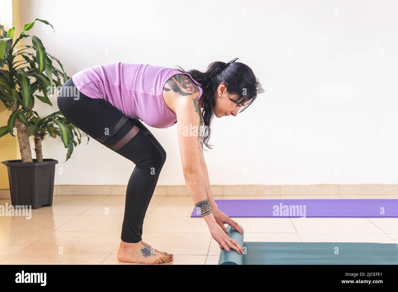 Donna latina adulta con tatuaggi che arrotolano un tappeto per la pratica dello yoga Foto Stock