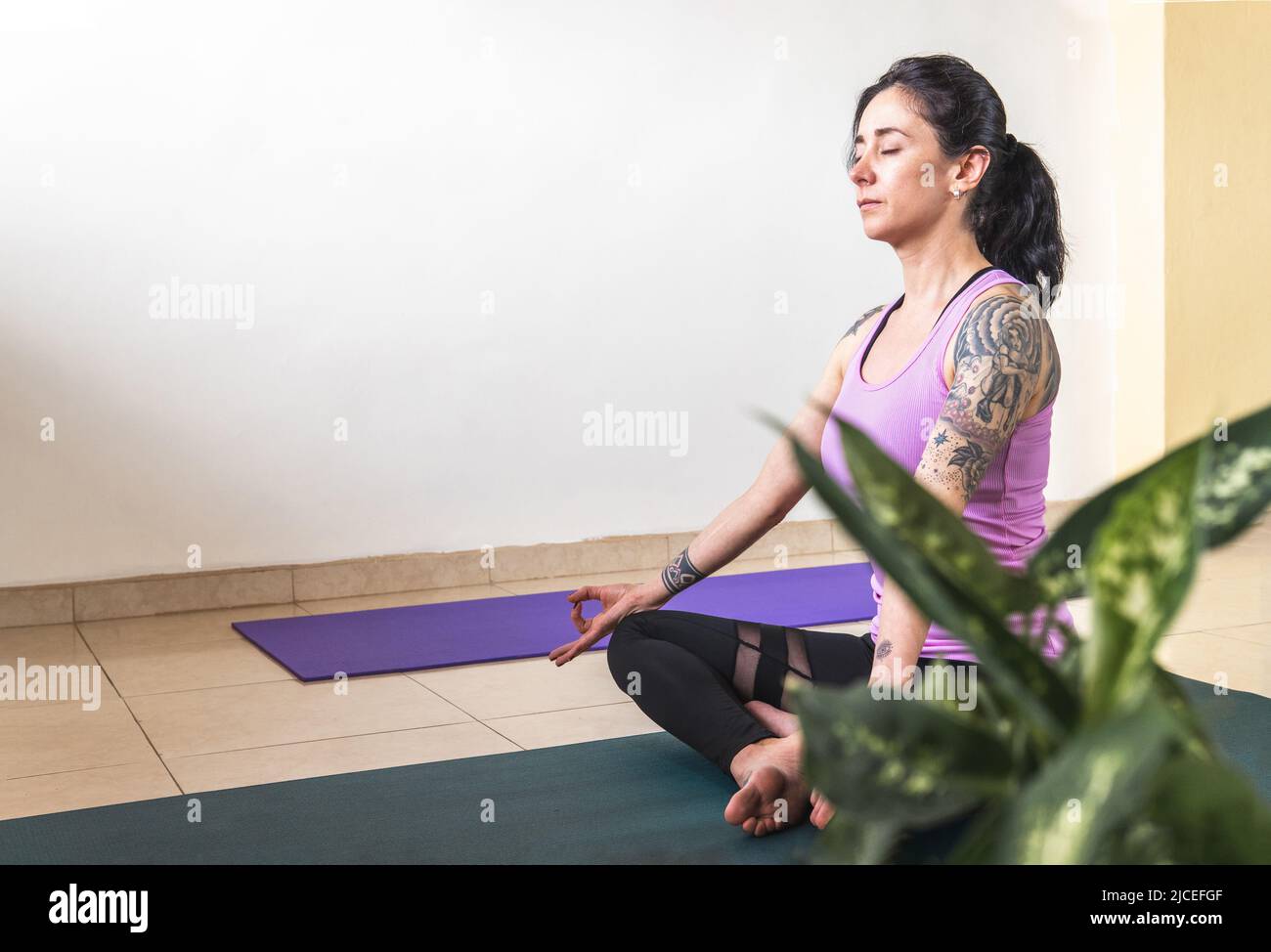 Donna latina dai capelli neri con tatuaggi che praticano lo yoga e la meditazione su un tappeto Foto Stock