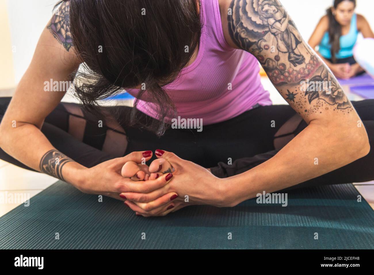 Primo piano focus selettivo della donna con tatuaggi in posizione Baddha Konasana o yoga Butterfly postura con un partner fuori fuoco dietro. Yoga e medi Foto Stock
