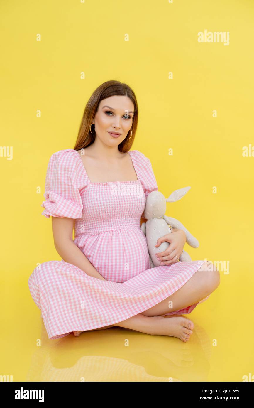 Donna con pancia in tarda gravidanza sedersi sul pavimento in abiti casual e tenere morbido giocattolo, sfondo giallo. Ritratto di giovane donna incinta attesa Foto Stock