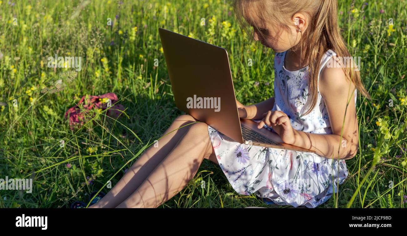 Bambina seduta sull'erba e tipi sulla tastiera del computer portatile. Istruzione, stile di vita, concetto tecnologico, concetto di apprendimento all'aperto Foto Stock