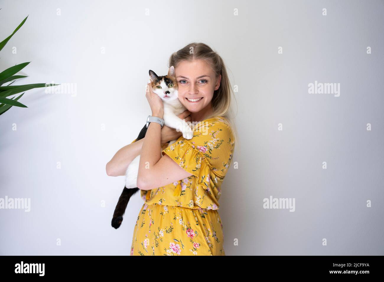 giovane donna bionda che porta il gatto bianco calico sulle braccia abbracciando il kitty sorridendo alla macchina fotografica guardando felice con lo spazio di copia Foto Stock