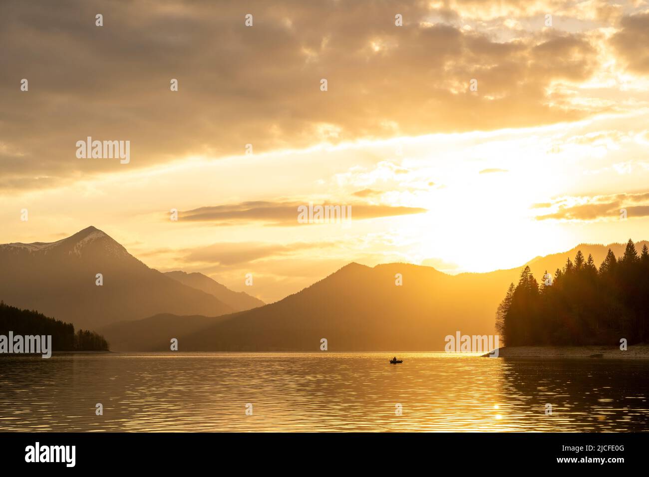 Atmosfera serale poco prima del tramonto a Walchensee nelle Alpi bavaresi, in primo piano un pescatore in una barca a remi, sullo sfondo il Simetsberg, l'atmosfera nuvolosa e il tramonto del sole. Foto Stock