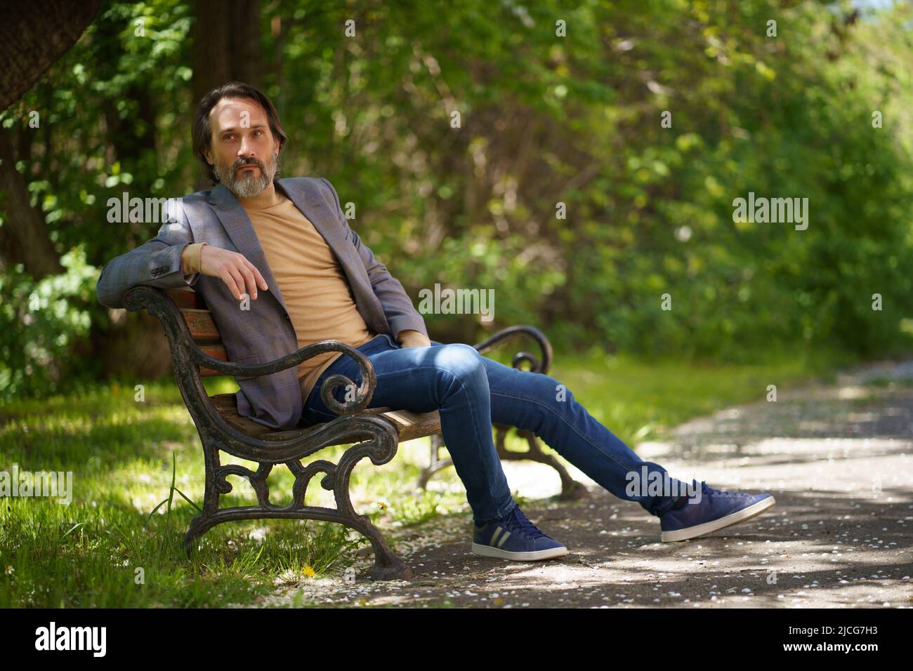 Uomo da freelance maturo con barba grigia, trascorri del tempo seduto sul panchina del parco godendoti del tempo libero o aspettando i colleghi indossando giacca e jeans casual. Foto Stock