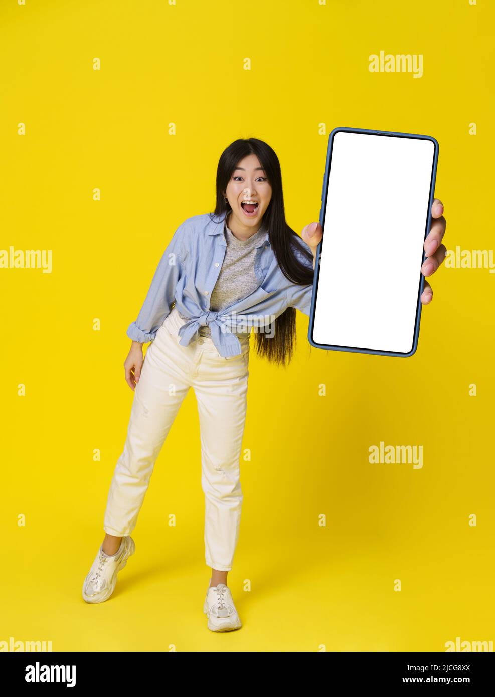 Giovane ragazza asiatica in casual happy holding smartphone mostra schermo bianco pubblicità app mobile e sorriso eccitato su fotocamera isolato su sfondo giallo. Grande offerta. Posizionamento del prodotto. Foto Stock