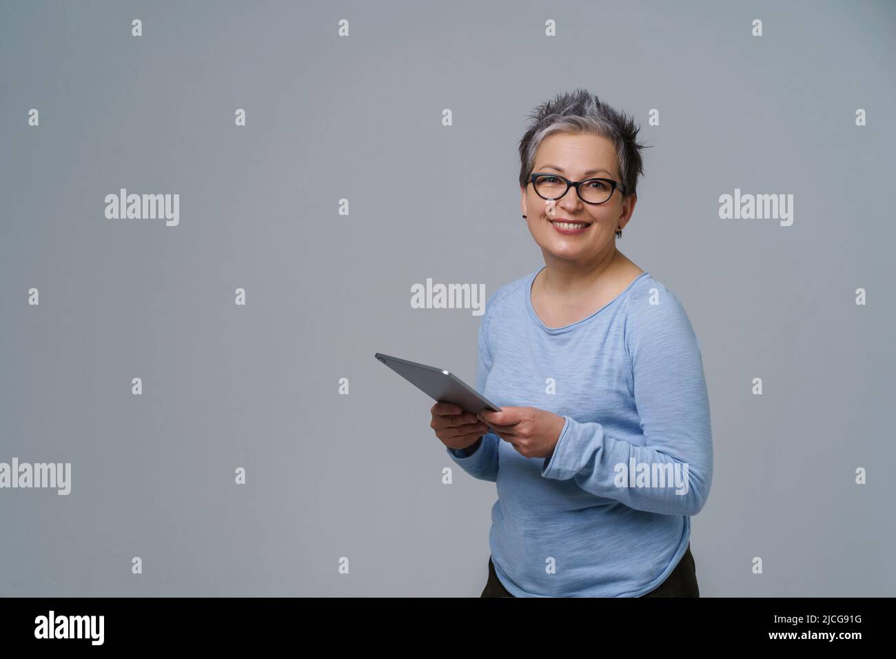 Donna d'affari in occhiali e tablet digitale in mani che lavora online sorridendo guardando la fotocamera. Bella donna nel 50s in blusa blu isolato su bianco. Persone anziane e tecnologie. Foto Stock