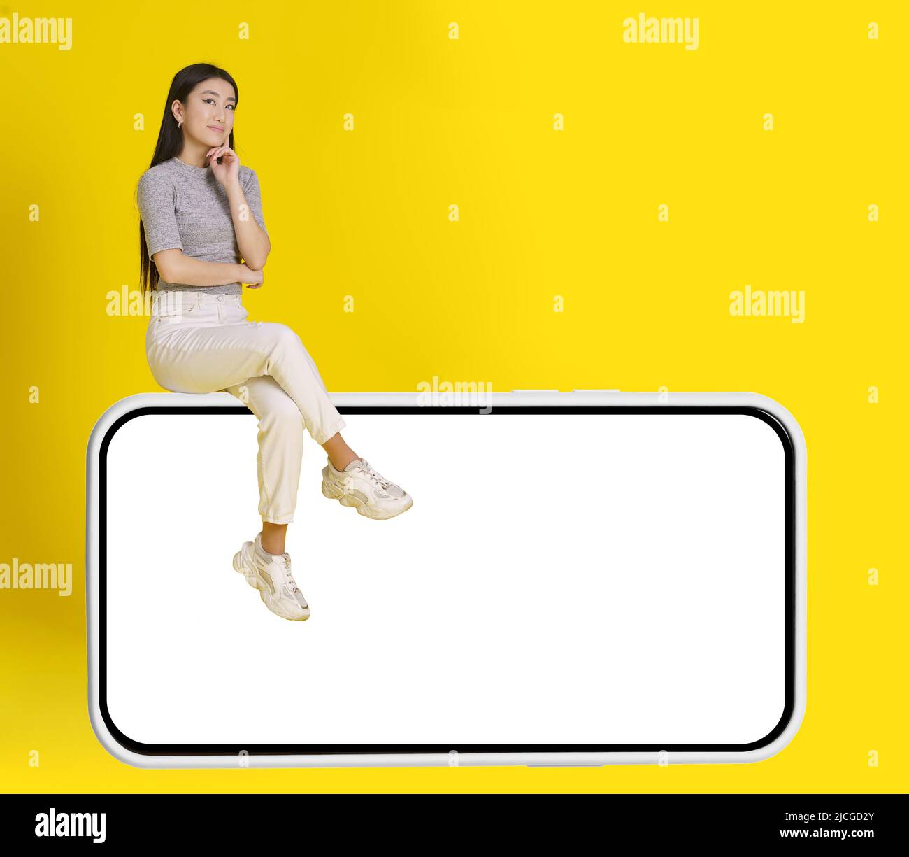 La ragazza asiatica pensierosa si siede su smartphone enorme con schermo bianco che indossa jeans bianco casual isolato su sfondo giallo. Simulazione del posizionamento dei prodotti. Pubblicità app mobile. Spazio di copia. Foto Stock
