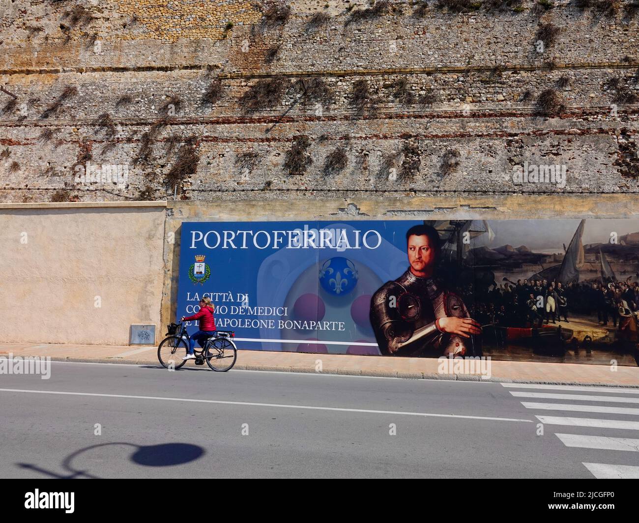 La donna corre in bicicletta davanti a un cartello, nella zona portuale di Portoferraio, Elba, pronunciando la città come luogo di Cosimo de Medici e Napoleone Bonaparte. Foto Stock