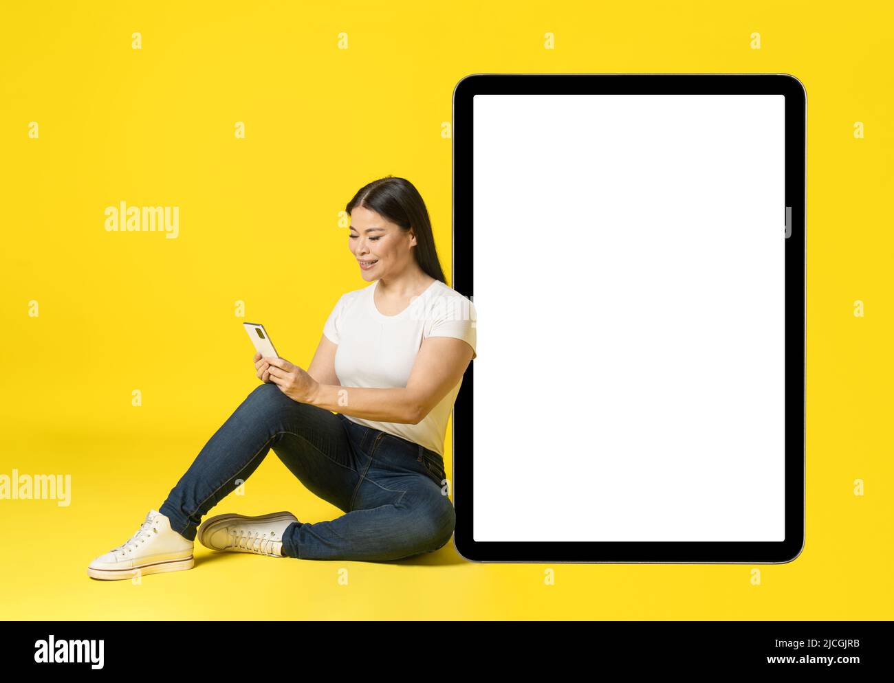Donna asiatica appoggiata su un enorme tablet PC gigante seduto sul pavimento con il telefono in mani in un casual bianco schermo, guardando la pubblicità cellulare app isolato sul giallo. Posizionamento del prodotto. Foto Stock