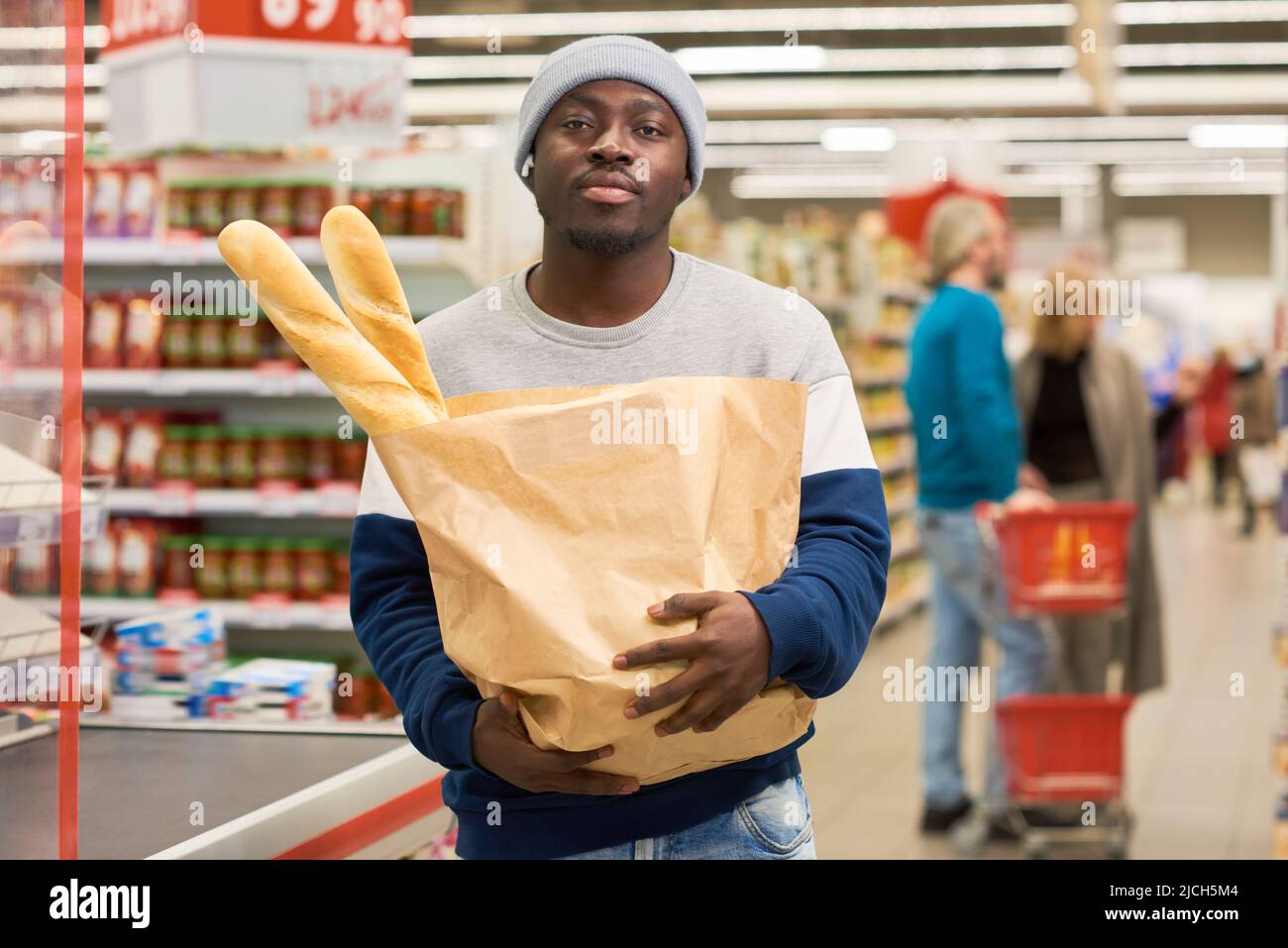 Giovane acquirente africano americano maschio che porta sacco di carta con due baguette di grano fresco e guardando la macchina fotografica contro altri clienti Foto Stock