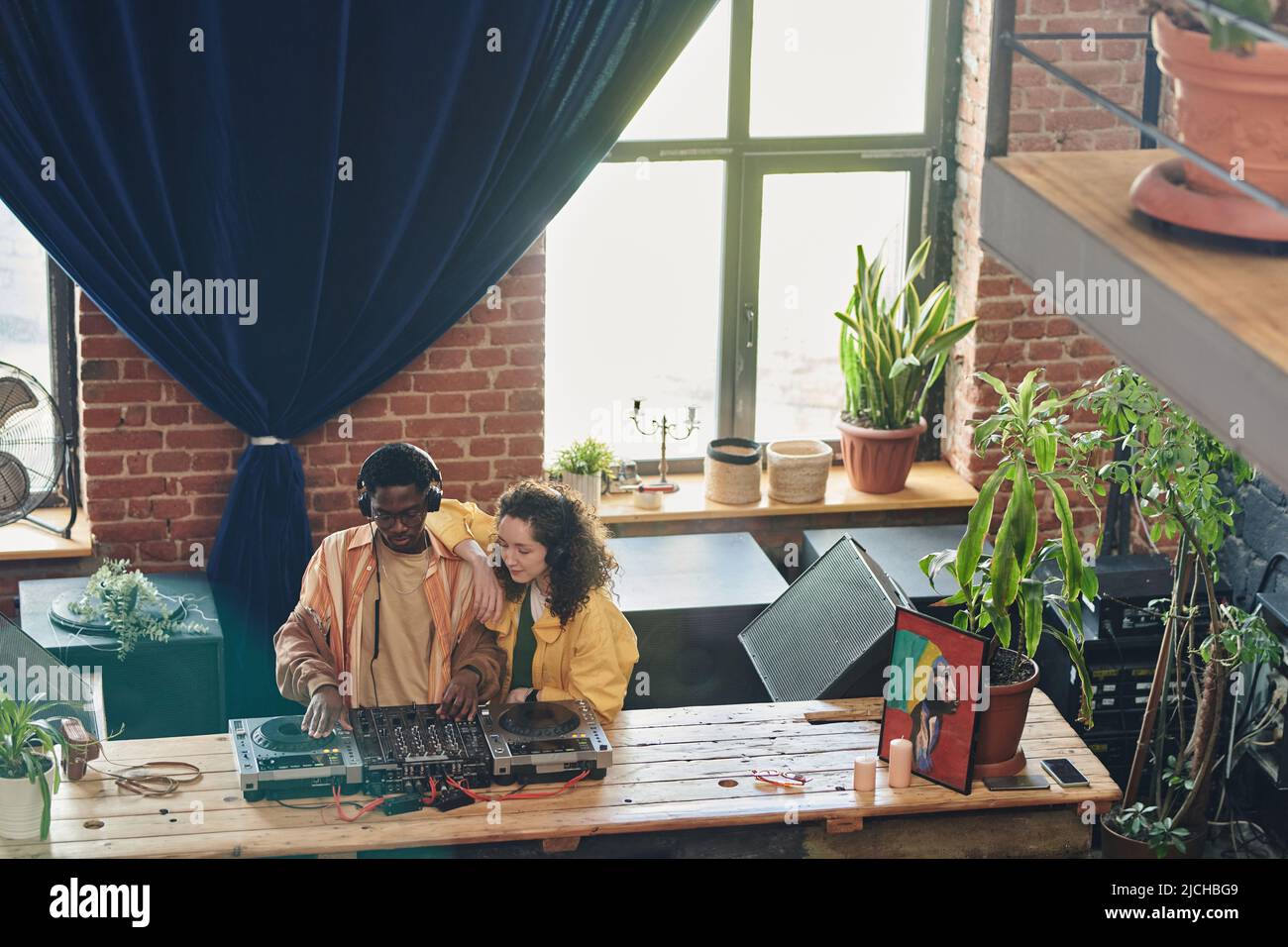 Ragazza con capelli ondulati in piedi vicino al giovane uomo nero mescolando suoni su stand dj mentre entrambi creano nuova musica da tavolo in appartamento loft Foto Stock