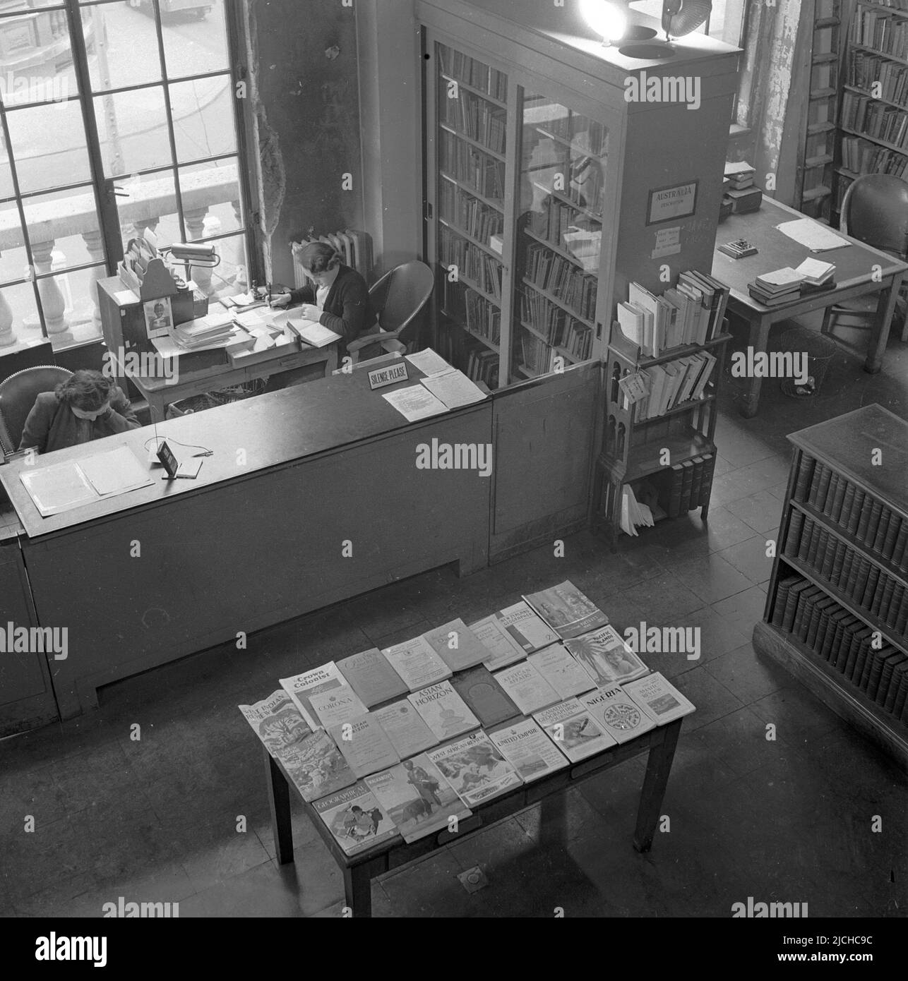 1950s, historicla, una vista dall'alto del banco di ricevimento ad una biblioteca pubblica con una signora che lavora ad una scrivania, Inghilterra, Regno Unito. Sul tavolo di fronte, sono esposti libri e opuscoli 'stranieri', tra cui la West African Review, United Empire, African Affairs. Foto Stock