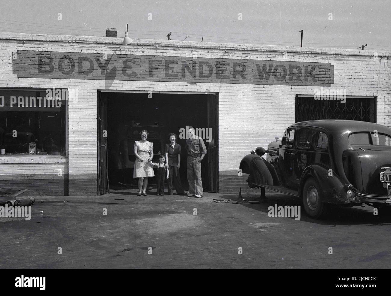 1930s, storica, due donne, un bambino piccolo e un'auto Body Worker in piedi per la loro foto all'entance ad un bodyworks auto in Utah, Mid-West, USA. Un cartello dipinto a mano sull'edificio dice: Carrozzeria e parafango, mentre un'automobile dell'epoca è parcheggiata all'esterno sul piazzale con la ruota anteriore sinistra rimossa, avendo fatto riparare e verniciare il parafango - che copre la ruota. Le vetture di questa epoca avevano parafanghi ampi e curvi che si estendeva dalla porta anteriore al paraurti anteriore. Foto Stock