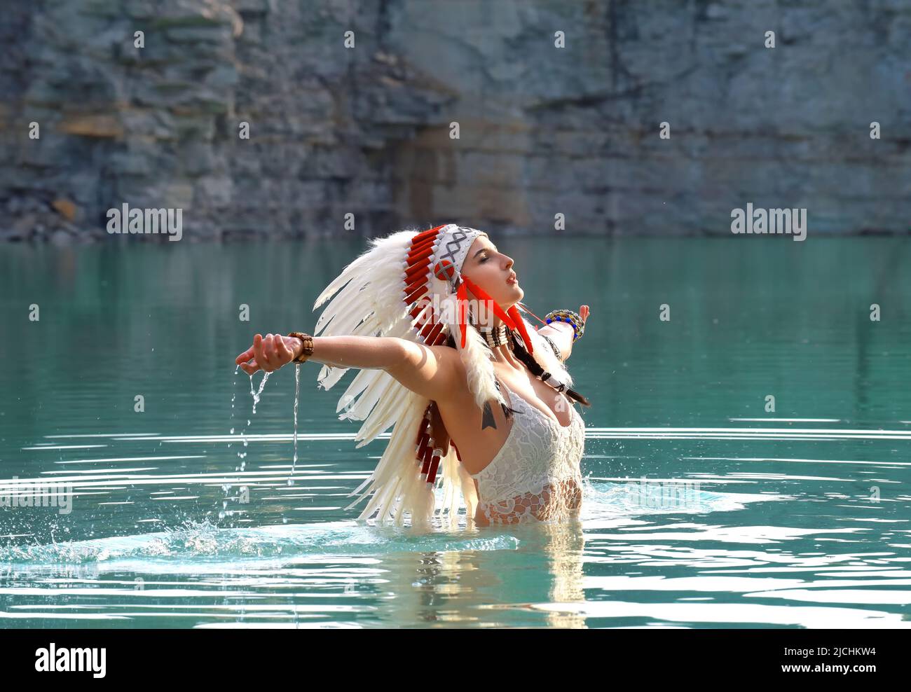 Una ragazza è vista vestita come un indiano nativo americano. Lei è vestita di bianco con un headdress piume. Si vede in piedi in un lago di cava. Foto Stock