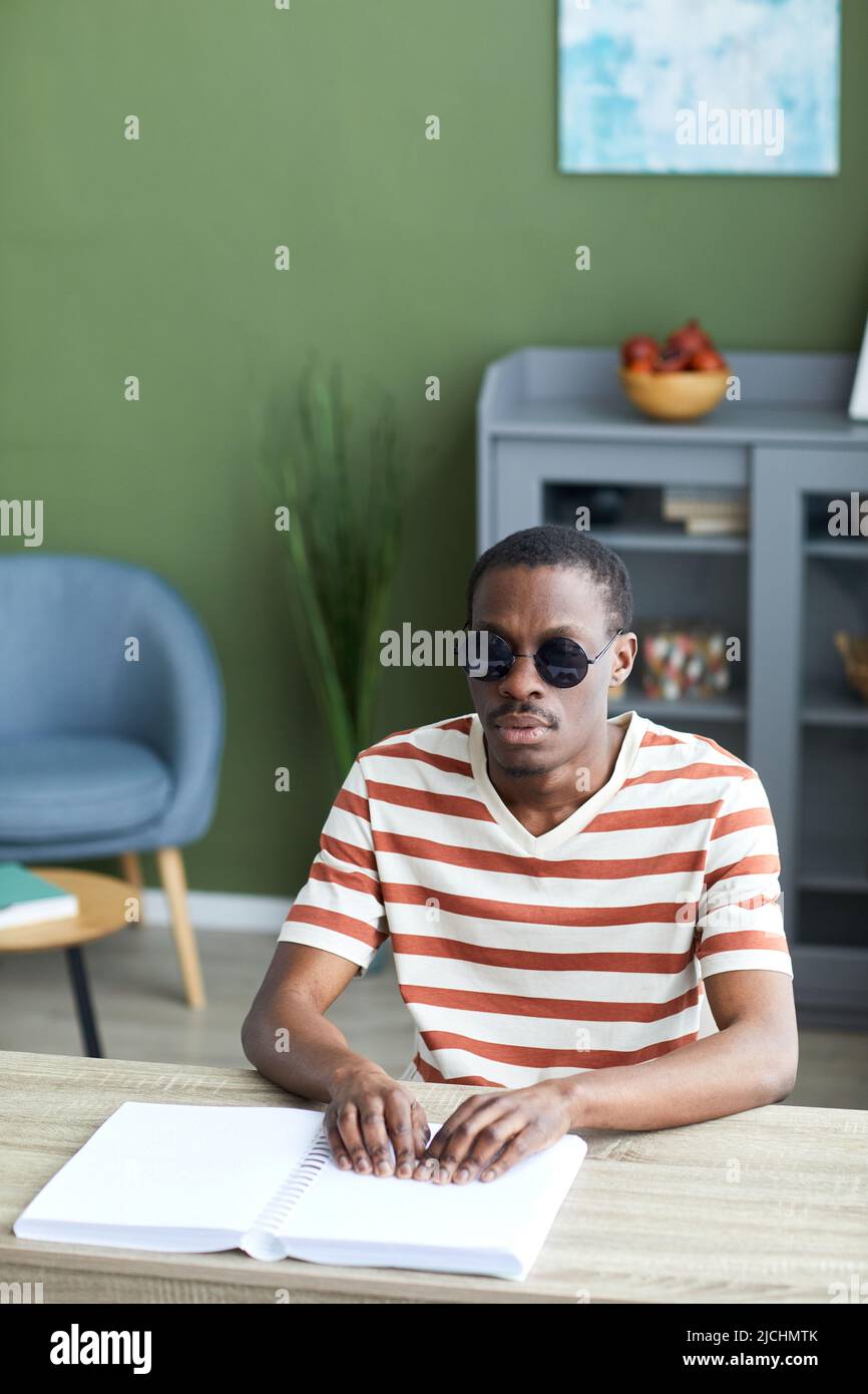 Ritratto verticale dell'uomo nero adulto che legge libro tattile in braille e indossa occhiali da sole scuri, informazioni accessibili concetto Foto Stock