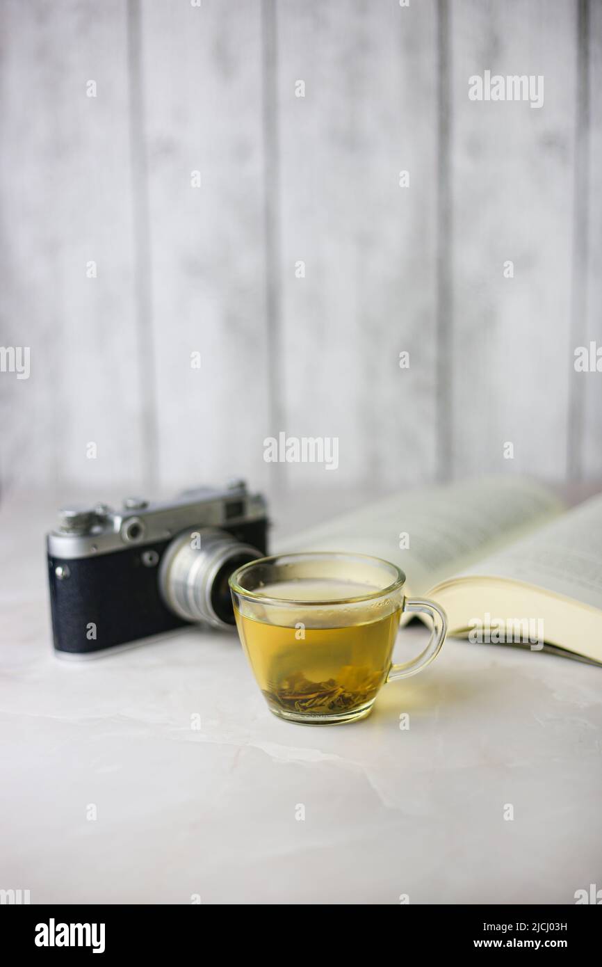 Tè verde alle erbe in una tazza trasparente accanto a un libro aperto e una macchina fotografica vintage su sfondo chiaro Foto Stock