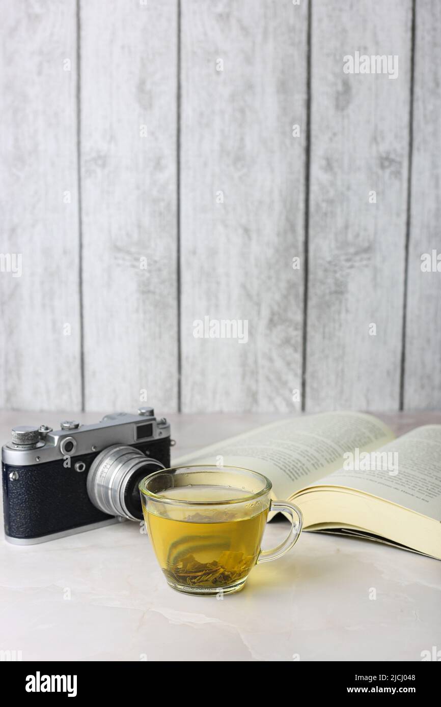 Tè verde alle erbe in una tazza trasparente accanto a un libro aperto e una macchina fotografica vintage su sfondo chiaro Foto Stock