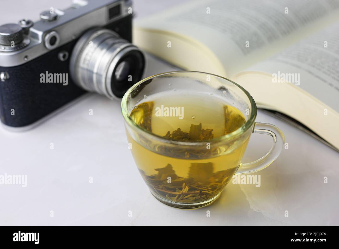 Tè verde alle erbe in una tazza trasparente accanto a un libro aperto e una macchina fotografica vintage su sfondo chiaro Foto Stock