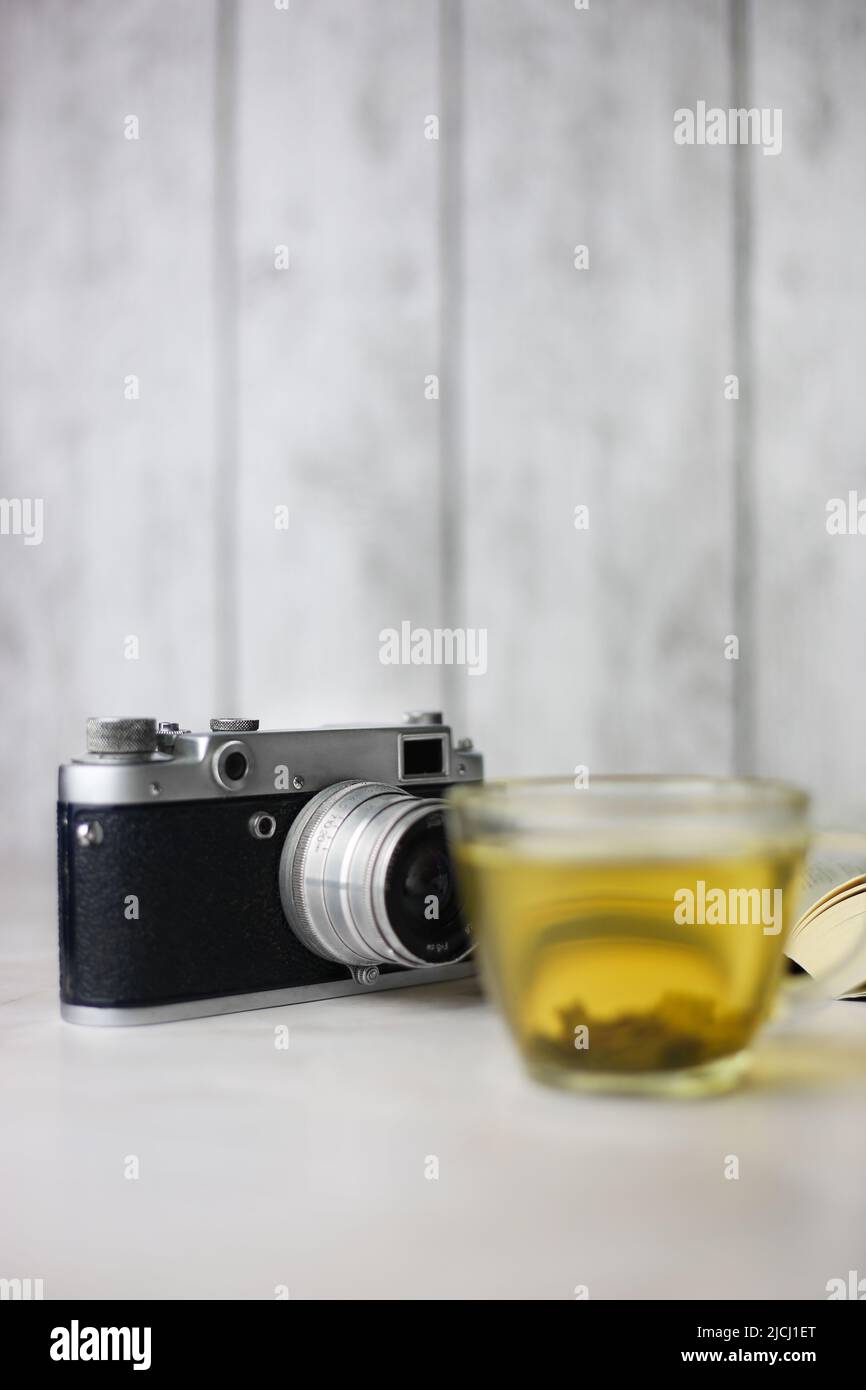 Tè verde alle erbe in una tazza trasparente accanto a un libro aperto e una macchina fotografica vintage su sfondo chiaro Foto Stock