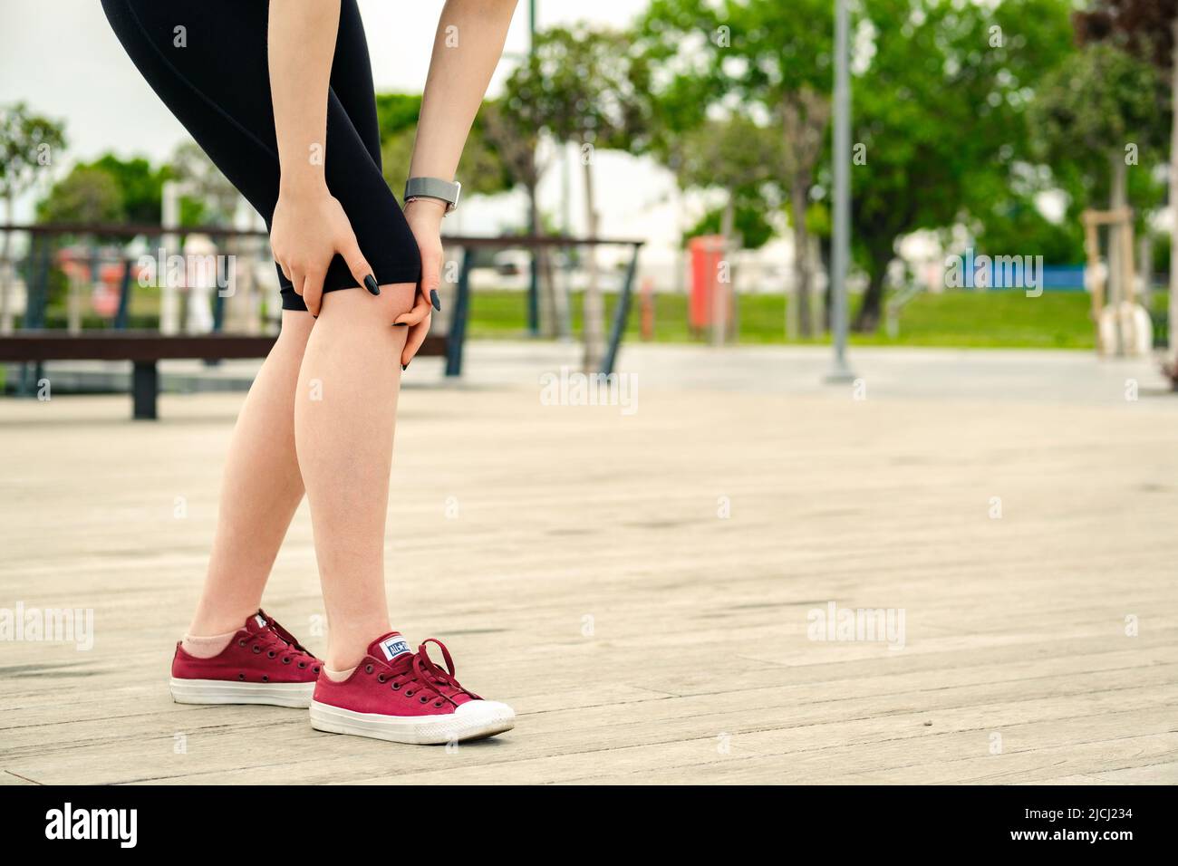 Giovane donna caucasica che indossa un reggiseno sportivo nero in piedi sul parco della città, all'aperto crampi alle gambe e sentire dolore, toccando la caviglia del ginocchio doloroso ferita fuori Foto Stock