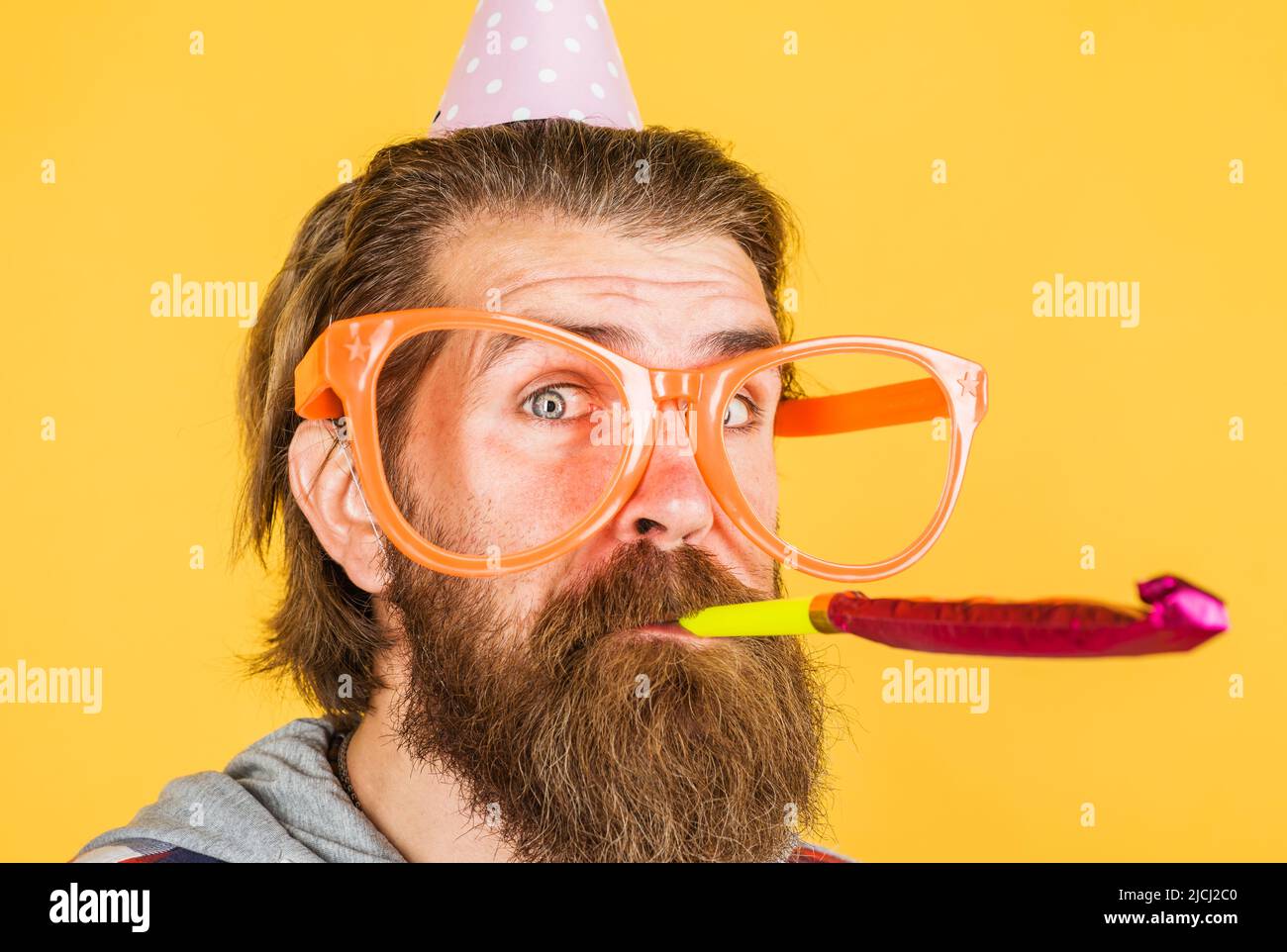 Uomo bearded che soffia il noisemaker. Festa di compleanno. Ora di festa. Ragazzo in cappello a cono e grandi bicchieri. Foto Stock