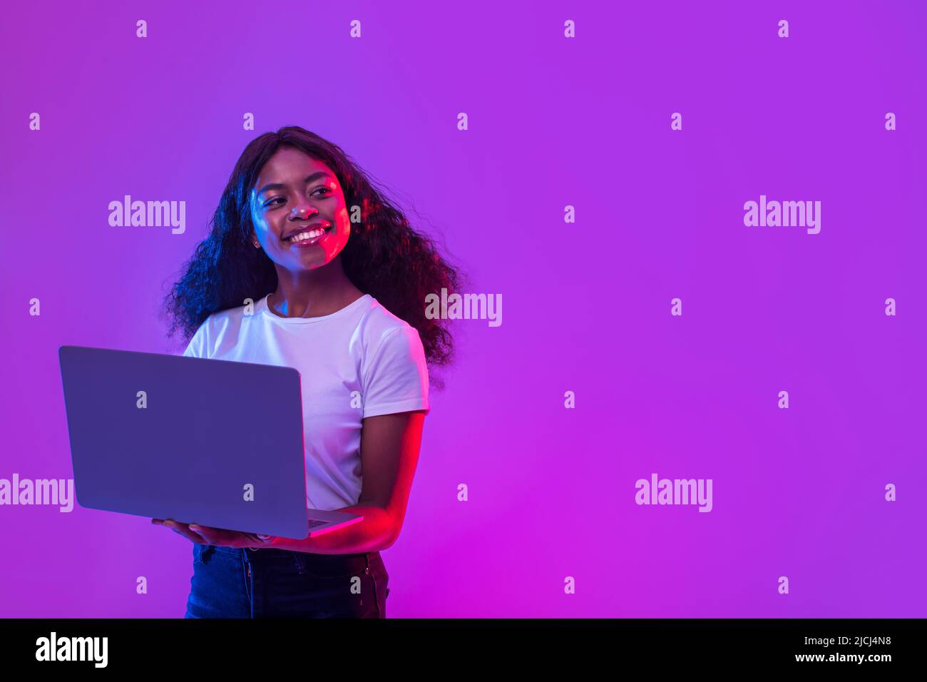 Signora afroamericana che usa il laptop per il lavoro in linea, l'istruzione o la comunicazione, guardando lo spazio vuoto nella luce del neon Foto Stock