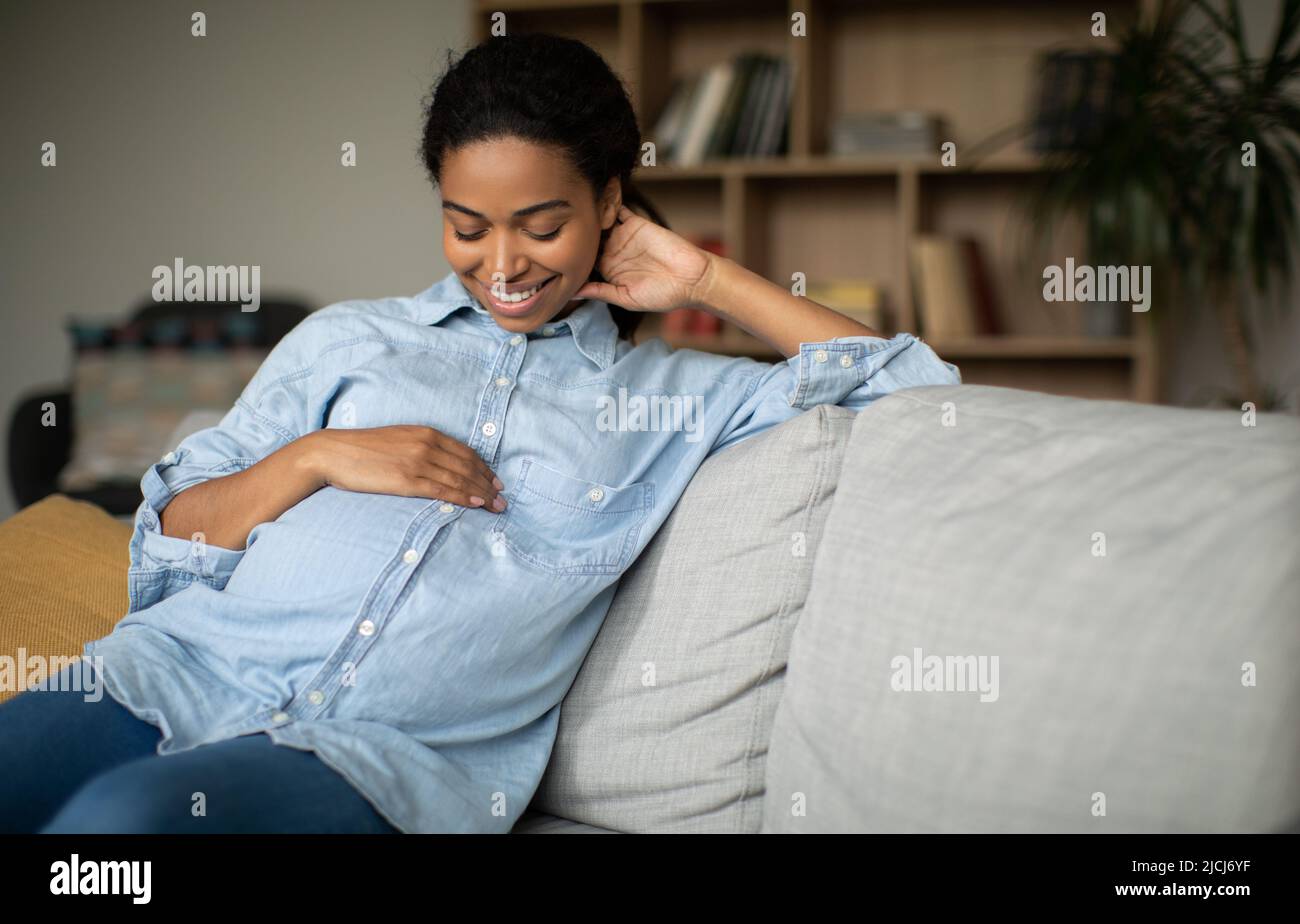 Happy incinta African American Woman sorridente stroking Belly seduta al coperto Foto Stock