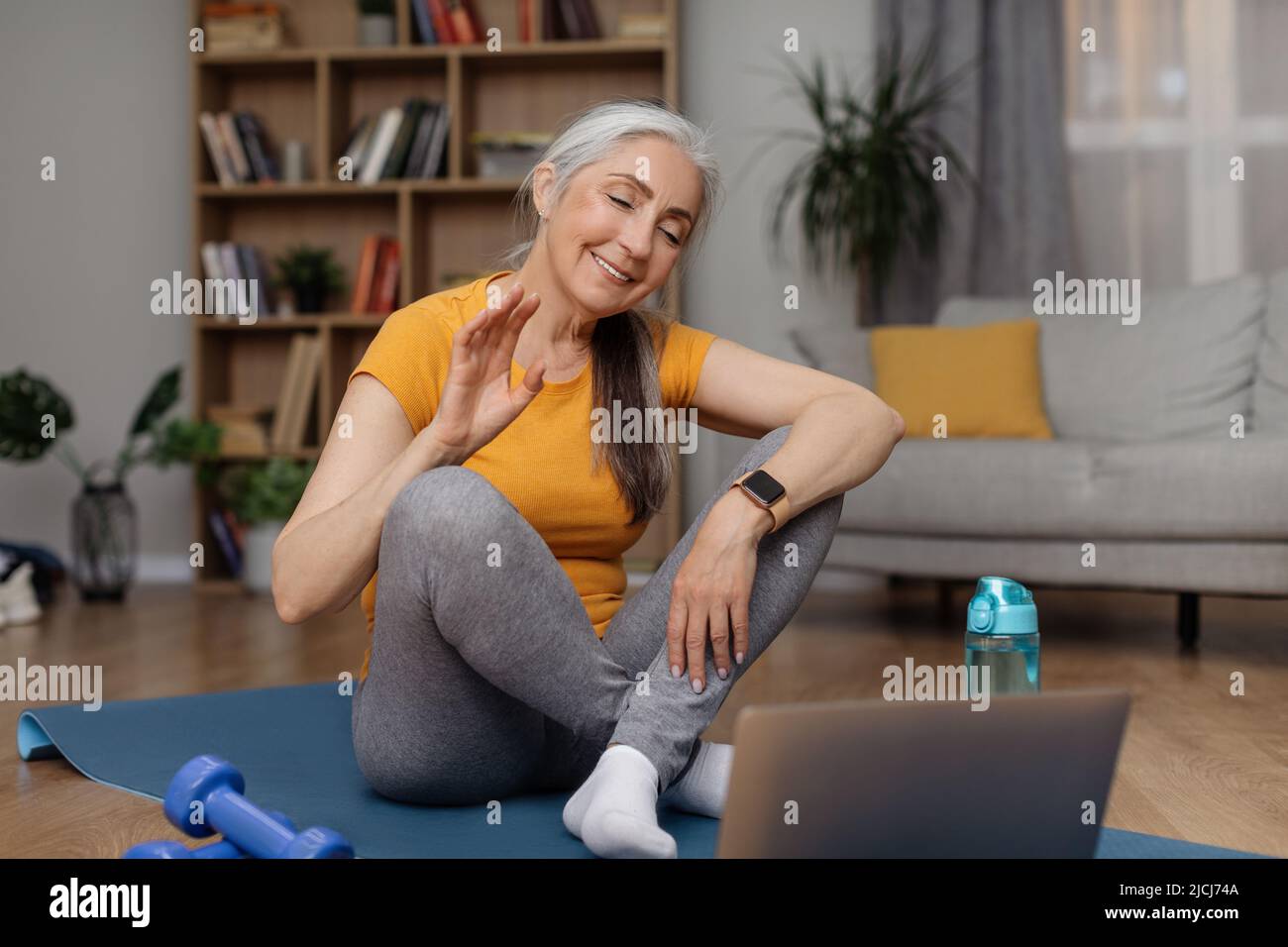 Donna anziana positiva che ondeggiava alla webcam del laptop facendo video conferenza con personal trainer, seduto sul tappeto yoga Foto Stock