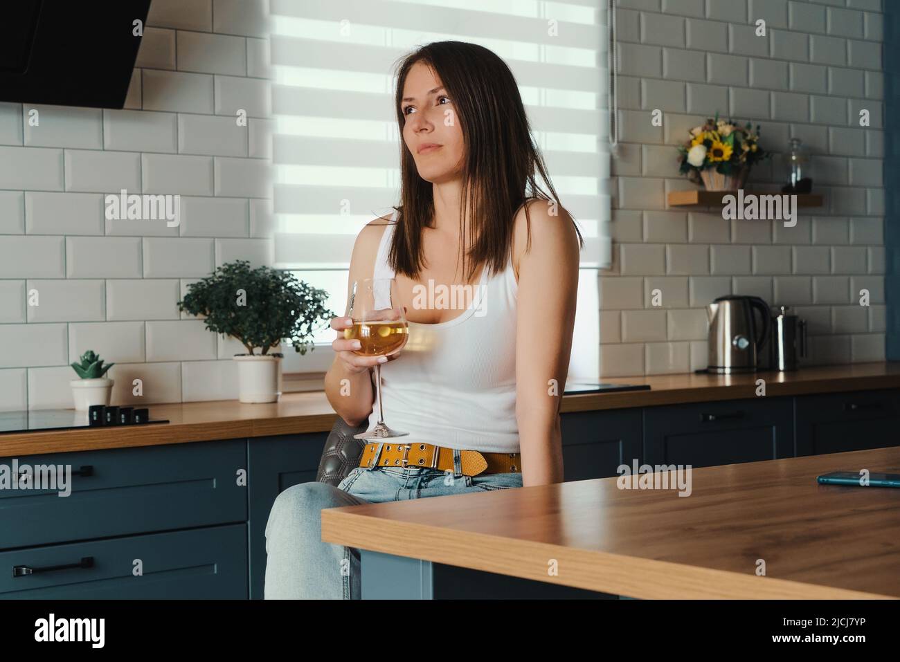 Giovane bella donna che tiene il bicchiere con vino bianco in mano seduto da solo in cucina a casa. Foto Stock