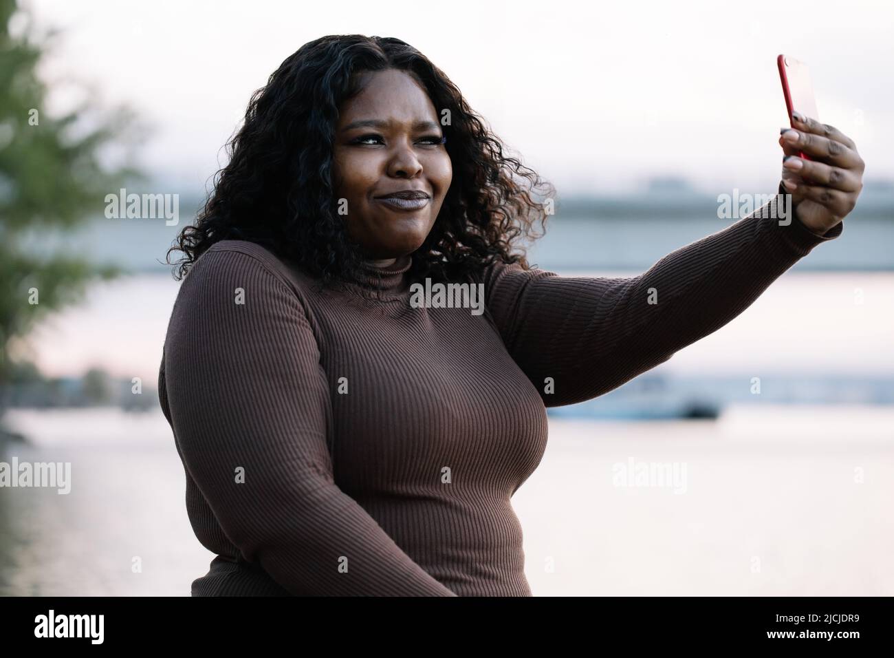 Curly fiducioso moderna multiculturale pelle scura donna in abito prendendo selfie al telefono, posa, grimacing. Corpo positivo Foto Stock