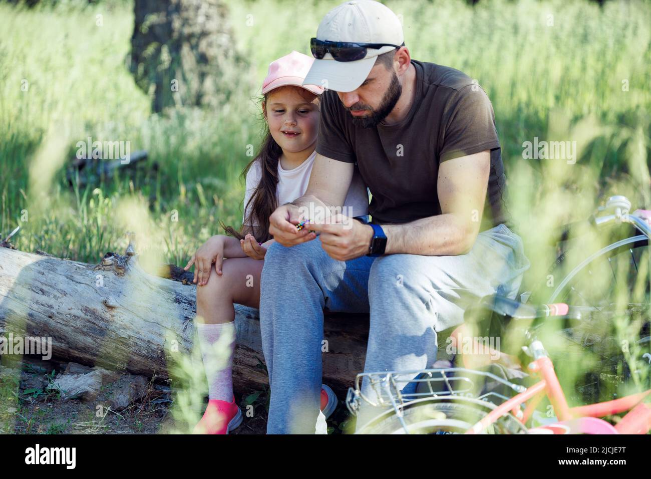 Ritratto di famiglia che indossa cappellini, seduta su asciutto log in parco. Piccola ragazza carina figlia che guarda le mani di uomo padre. Foto Stock