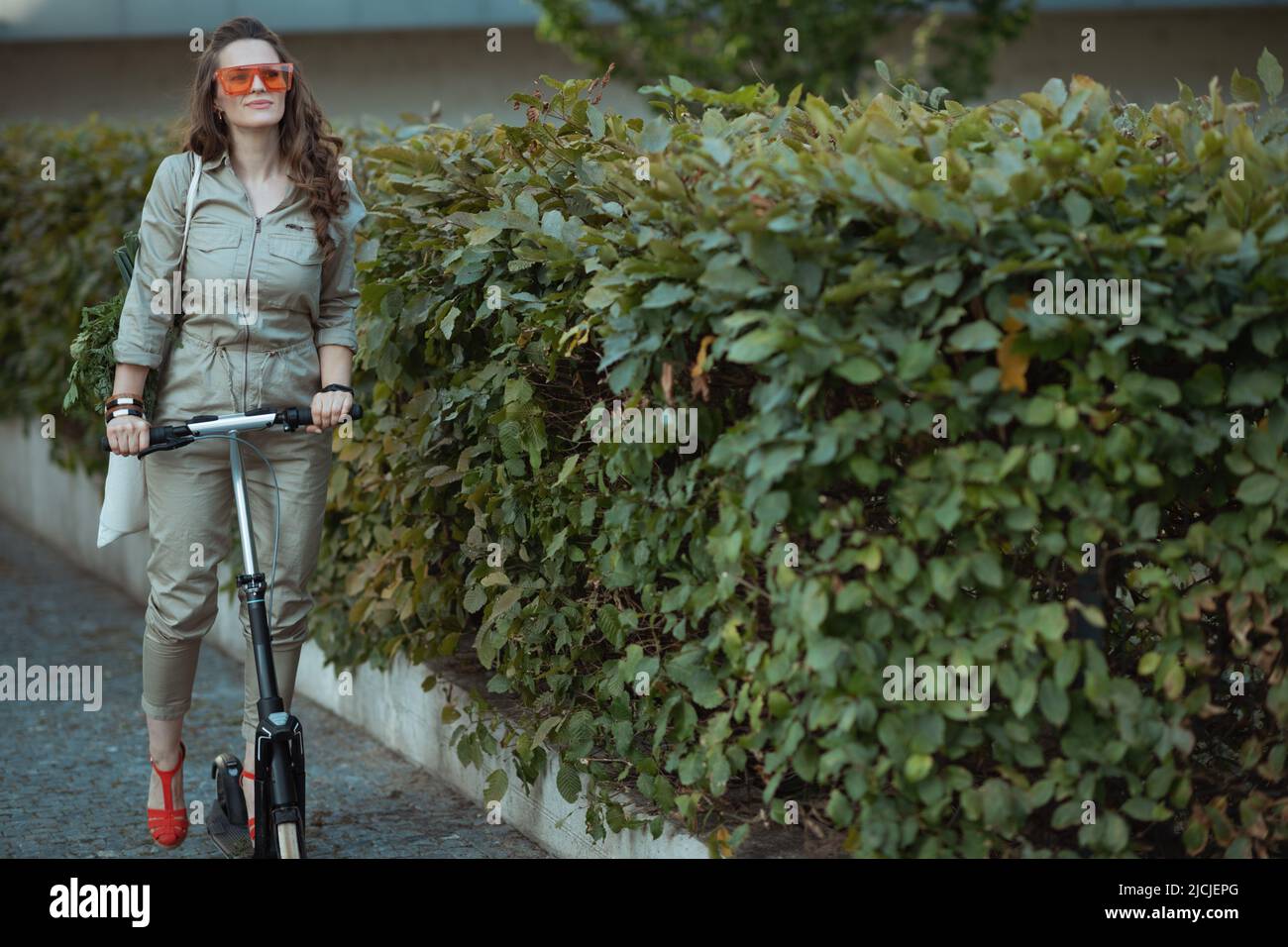 donna moderna in occhiali da sole e generale con borsa tote e scooter vicino recinto verde fuori della città. Foto Stock