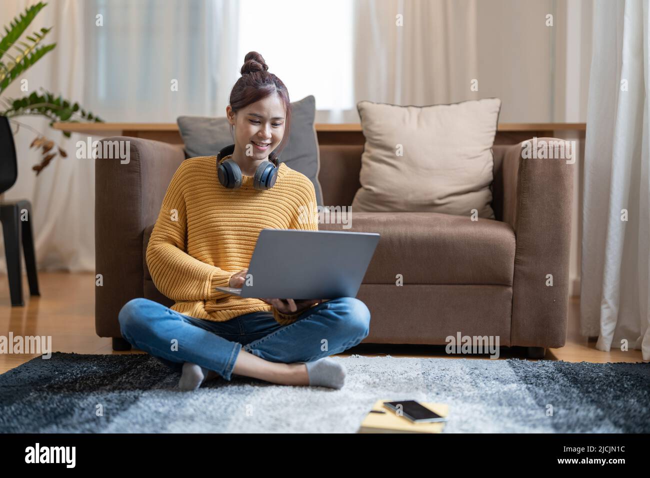 Giovane donna asiatica vestita casualmente lavorando su un computer portatile mentre si siede sul pavimento a casa. Lavorate da casa in un'atmosfera accogliente Foto Stock