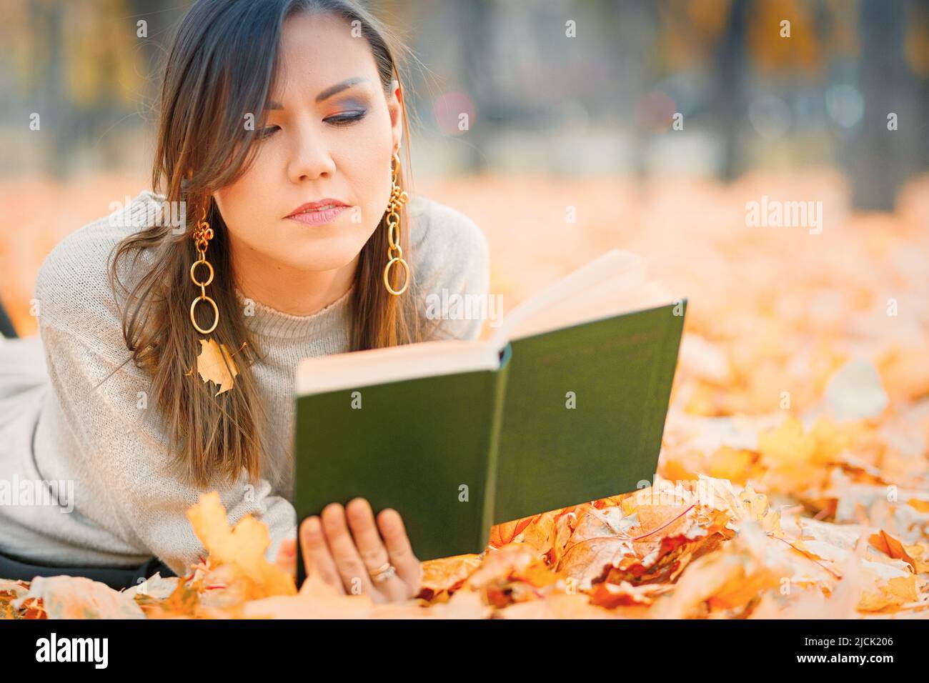 Donna dell'asia centrale sdraiata sul fogliame autunnale e libro di lettura nel parco Foto Stock
