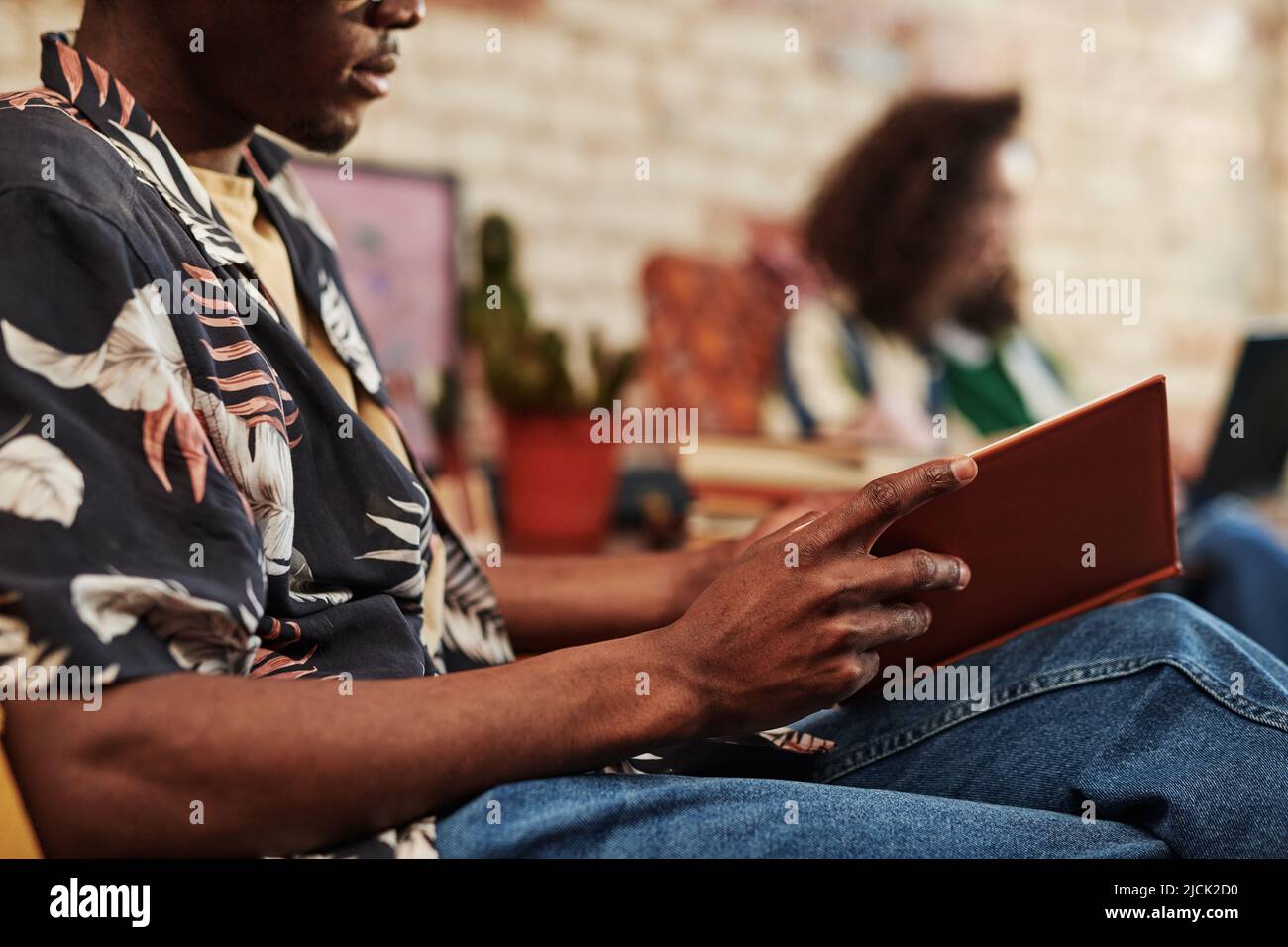 Mani di giovane uomo nero in jeans e camicia apertura libro in copertina marrone dura mentre seduto in poltrona a piacere contro la sua ragazza Foto Stock