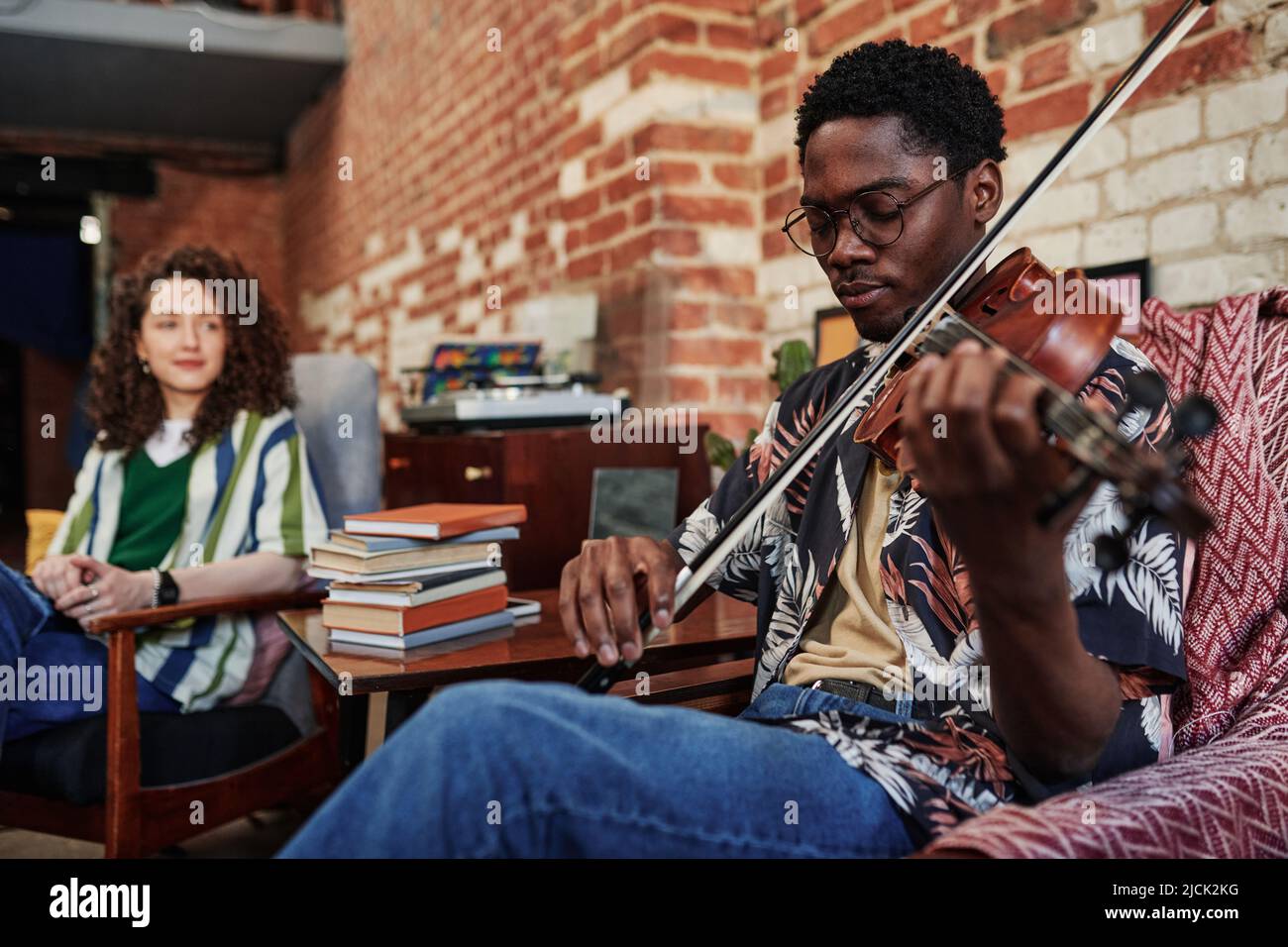 Giovane uomo in occhiali e casualwear seduto in poltrona in soggiorno di appartamento loft a piacere e giocare violino per la sua ragazza Foto Stock