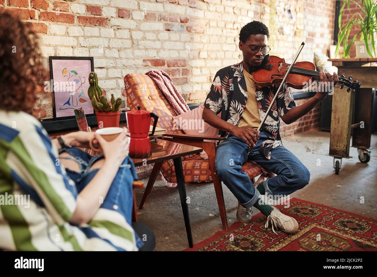 Guy in casualwear di 90s suonando violino mentre si siede in poltrona davanti alla sua ragazza che ha il tè e ascoltando un pezzo di musica classica Foto Stock