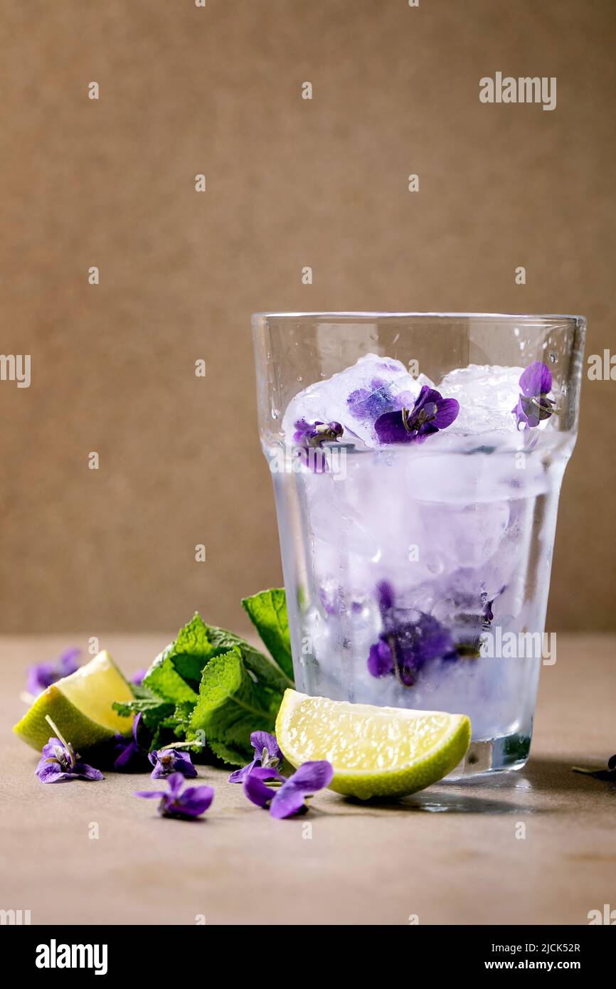 Bicchiere trasparente di cocktail soda con limonata floreale e menta, violetti, ghiaccio di fiori e fettine di lime in piedi sul tavolo marrone. Freddo drin estate rinfresco Foto Stock