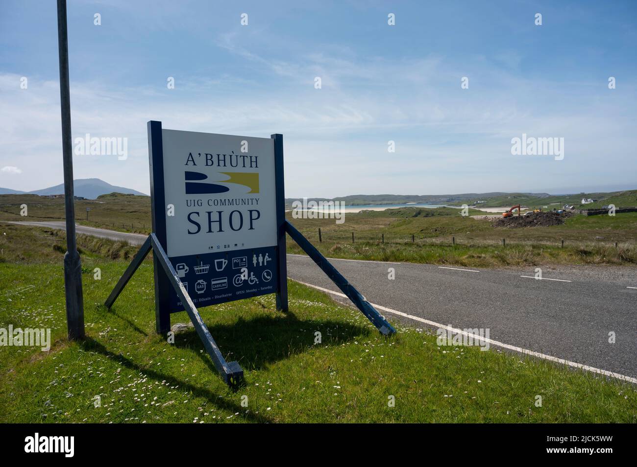 Insegna per il negozio della comunità Uig sull'isola di Lewis con icone per carburante, shopping, rinfreschi ecc. strada, campi, spiaggia e mare in background. Giorno di sole Foto Stock