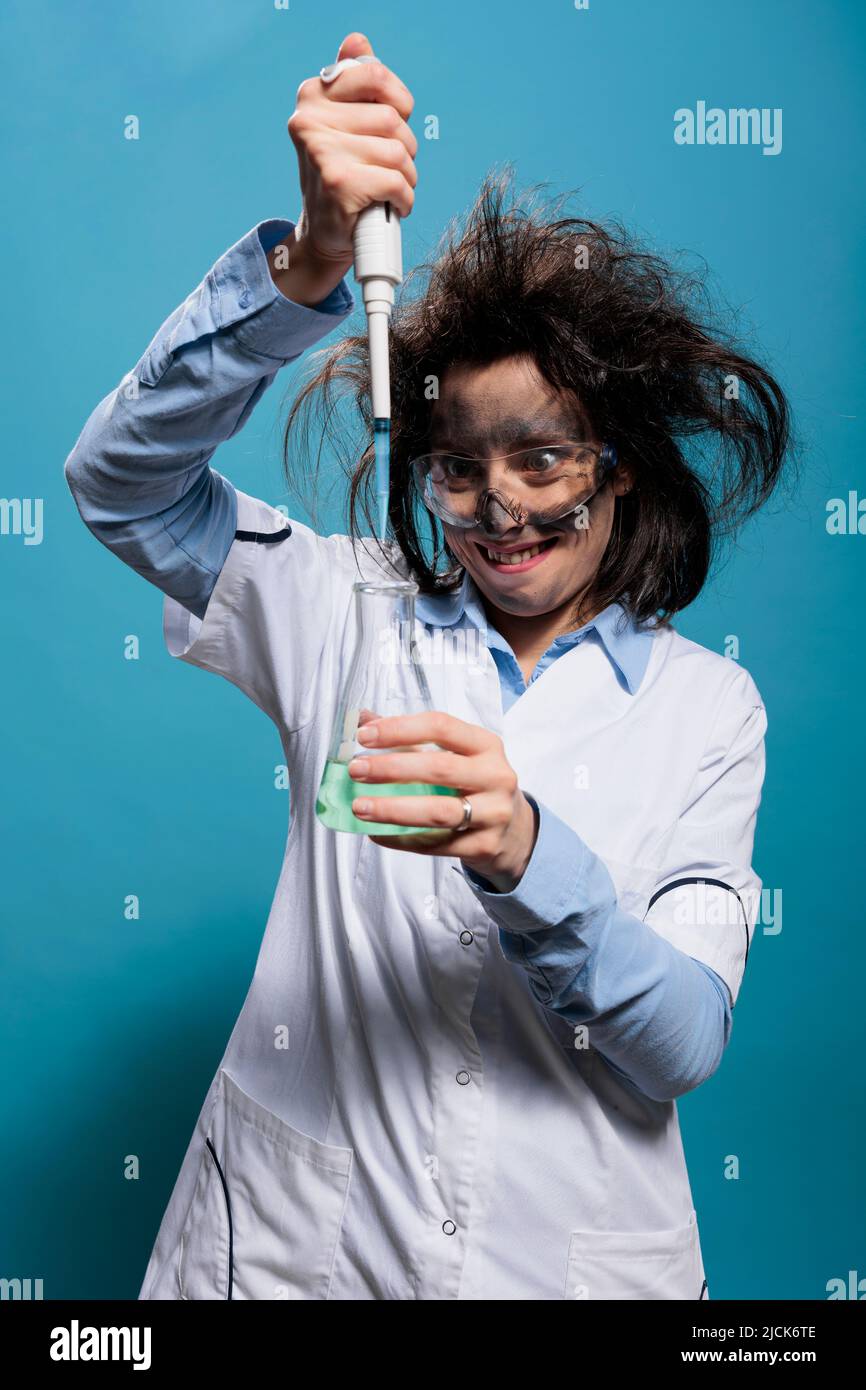 MAD sciocco lavoratore di laboratorio utilizzando la pipetta per mescolare siero sperimentale mentre grinning terribile. Scienziato insano mentre gocciola i composti chimici dalla pipetta in un pallone di vetro riempito con sostanza liquida. Foto Stock