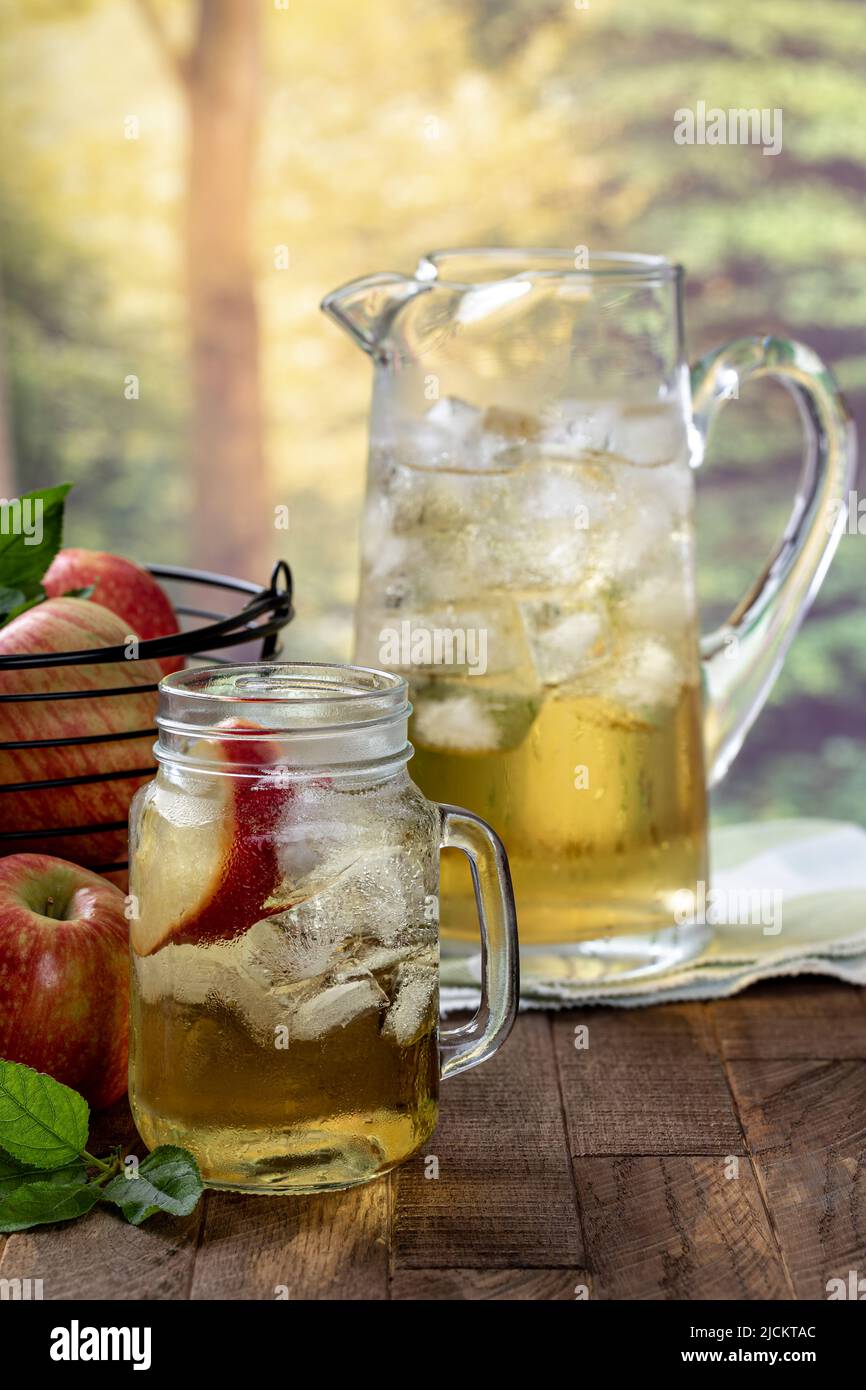 Bicchiere di succo di mela con fetta di mela e ghiaccio e un cestino di mele e caraffa su un tavolo di legno con sfondo rurale estivo Foto Stock