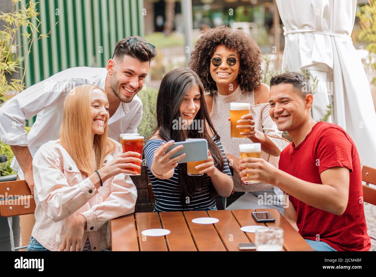 Gruppo di giovani amici, uomini e donne felici e diversi, prendendo selfie di gruppo, mentre si ha una birra al tavolo del pub all'aperto Foto Stock