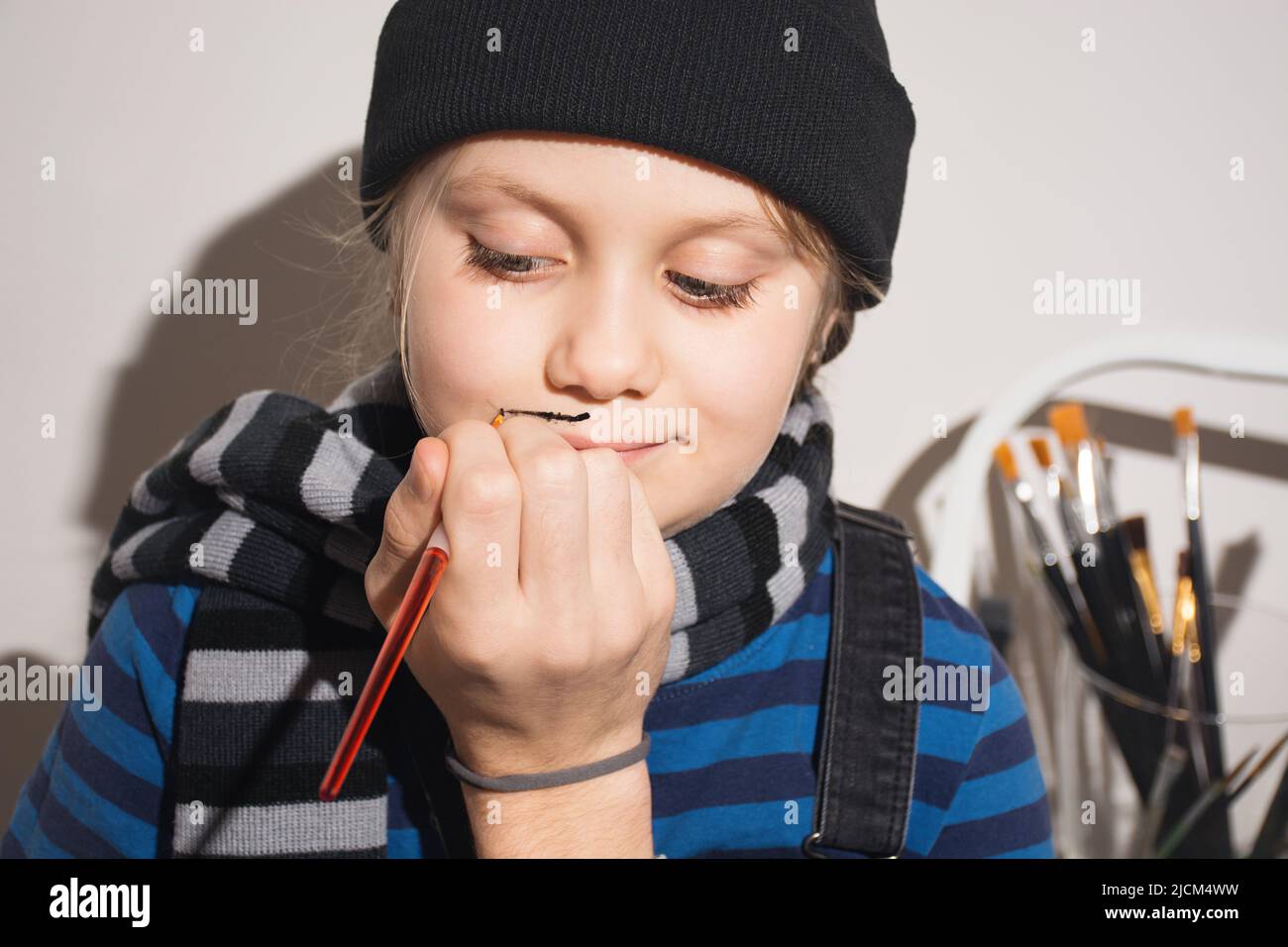 Primo piano ritratto ragazza. I bambini dipingono con il pennello sulle loro facce baffi francesi neri puliti per Halloween, mascherate di Capodanno, compleanno. Cappello e sciarpa su ragazza. Faccia arte. Persone reali. Spazio di copia Foto Stock