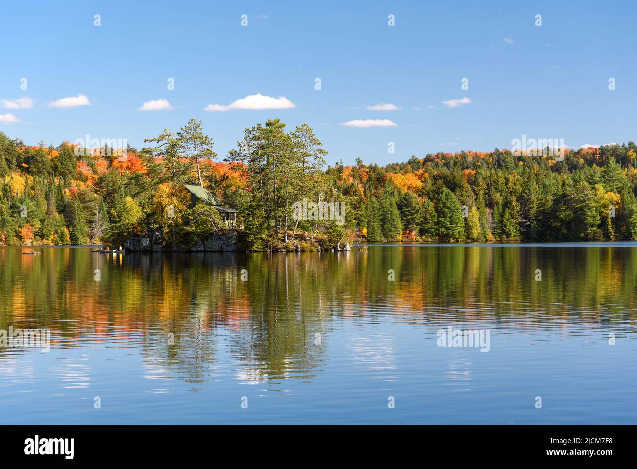Cabina su una piccola isola rocciosa nel mezzo di un lago circondato da fitta foresta in una soleggiata giornata autunnale. La gente in canoa si trova nei pressi dell'isola. Foto Stock