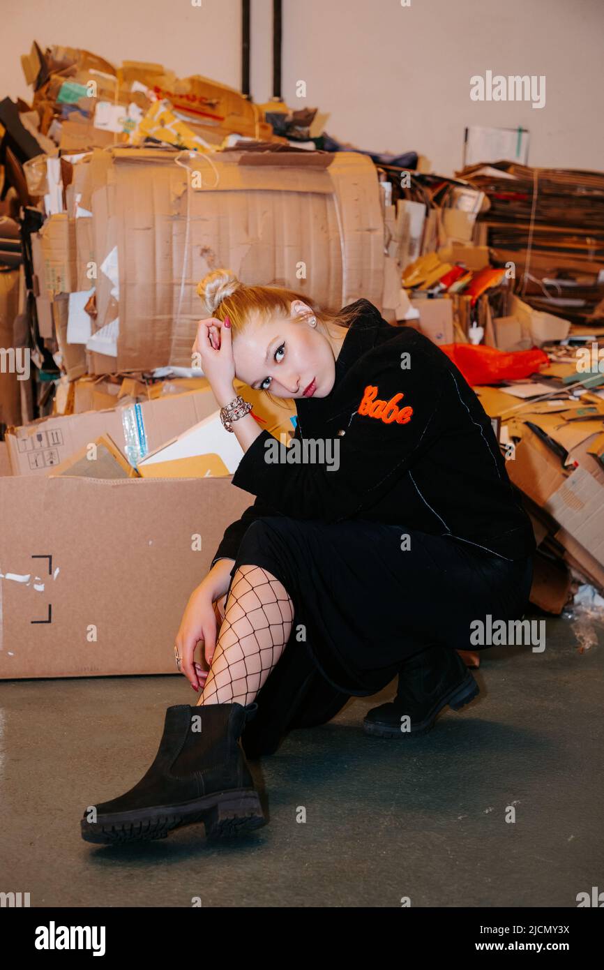 Ragazza bionda graziosa con bun dei capelli, portando un cappotto nero lungo, calze rete & stivali neri, siede vicino contenitore dei rifiuti con carta & cartone; riciclaggio? Foto Stock